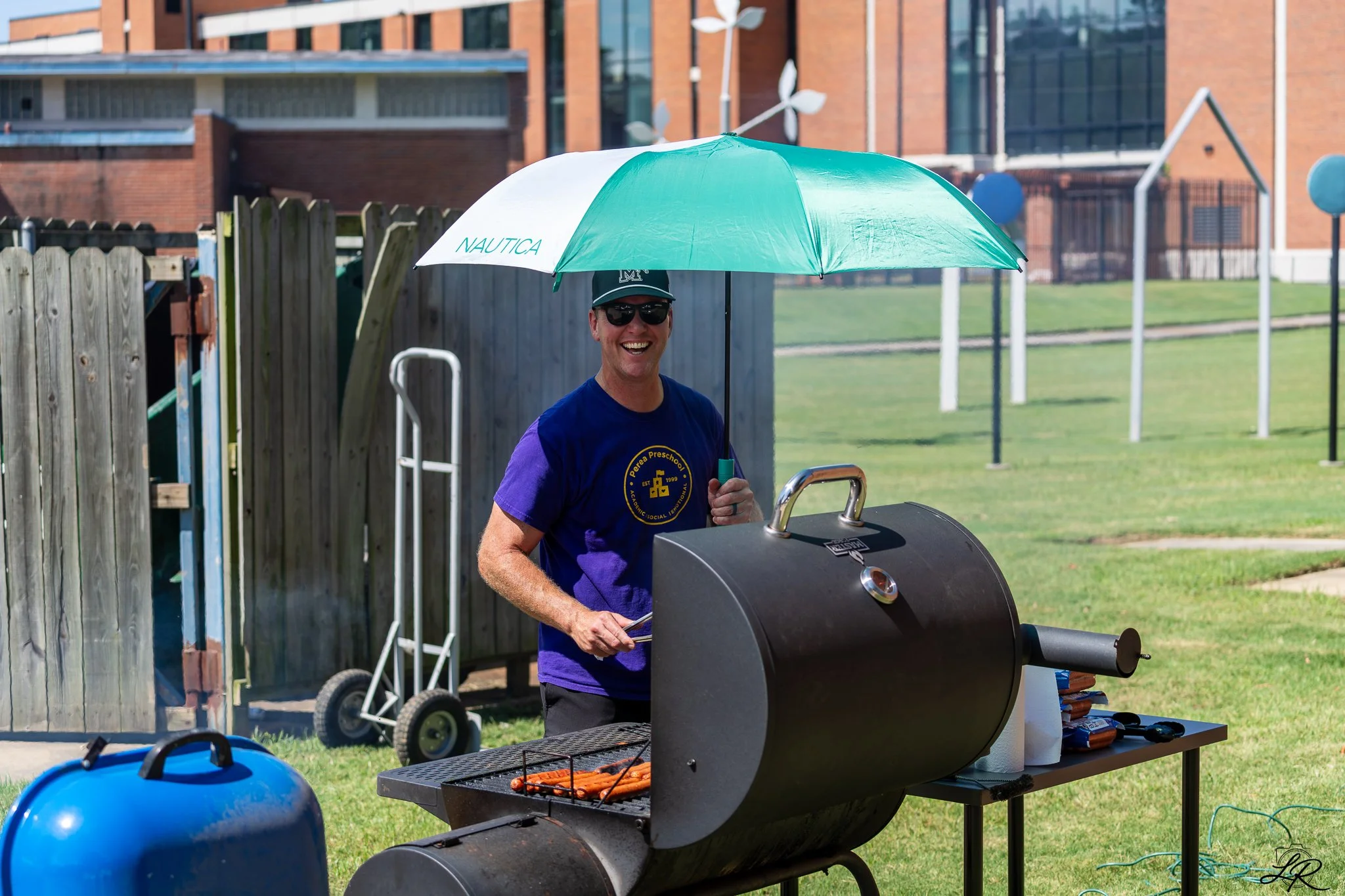 Man grilling hot dogs on a barbecue during an outdoor event on a sunny day, smiling, wearing sunglasses, a cap, and a navy blue t-shirt.