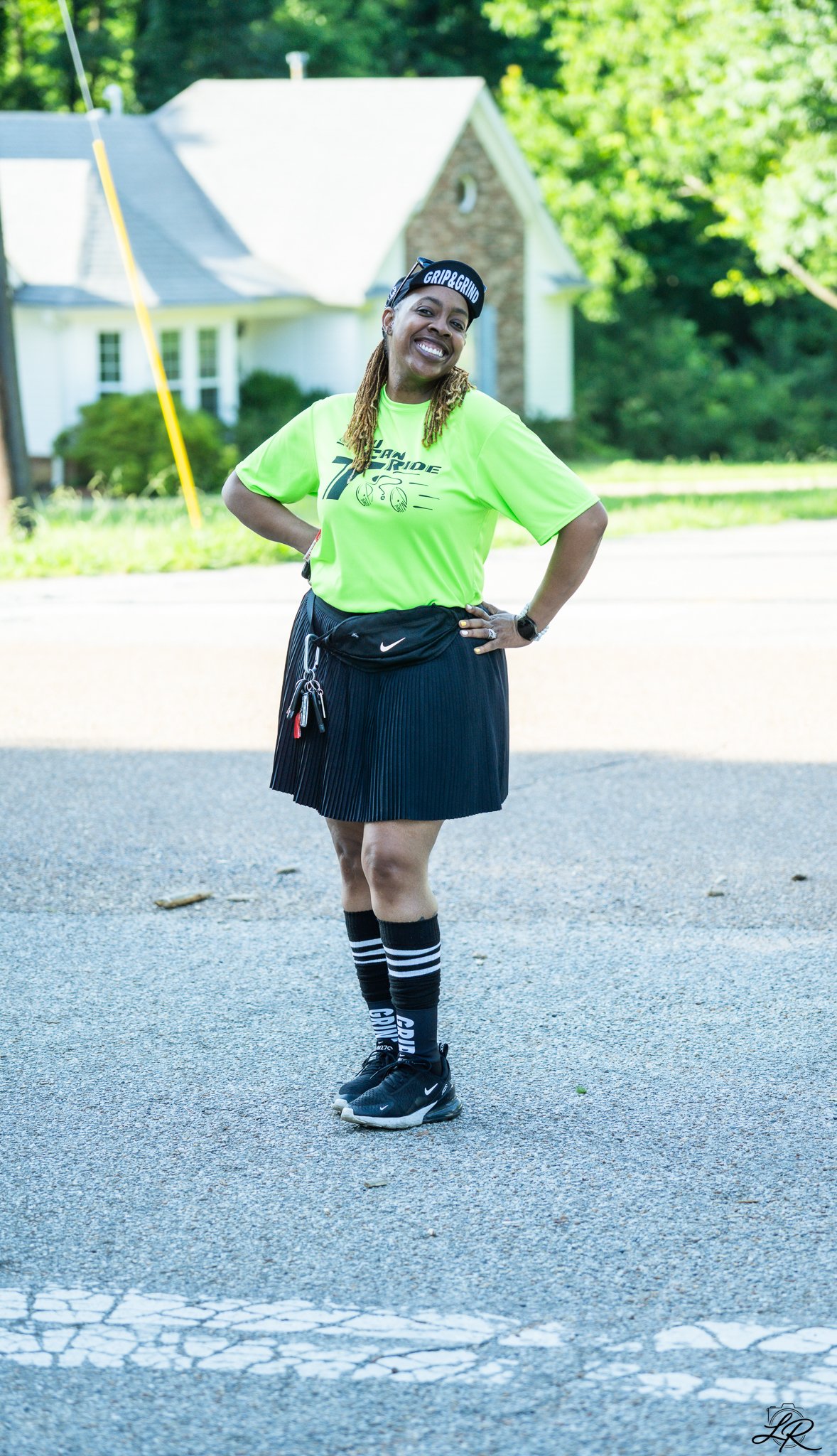 A woman is standing on a street with her hands on her hips, smiling. She wears a bright yellow T-shirt, a black pleated skirt, knee-high black socks with white stripes, and black Nike sneakers. She has a black waist pouch and a black visor with white