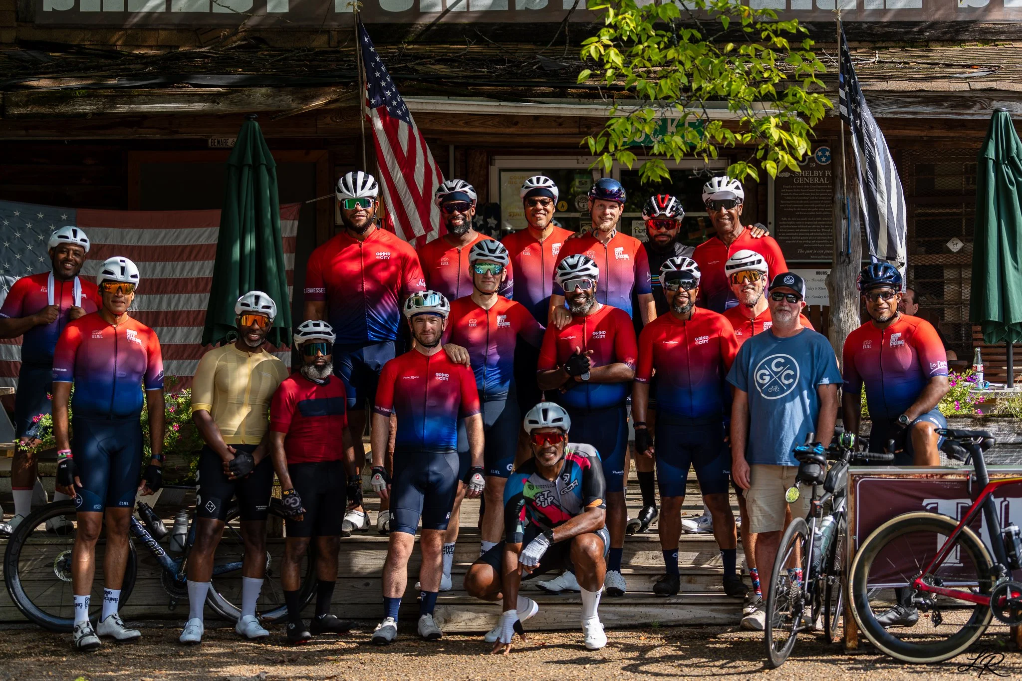 A group of cyclists in colorful cycling gear and helmets, posing in front of a wooden building with American flags and green umbrellas, outdoors on a sunny day.