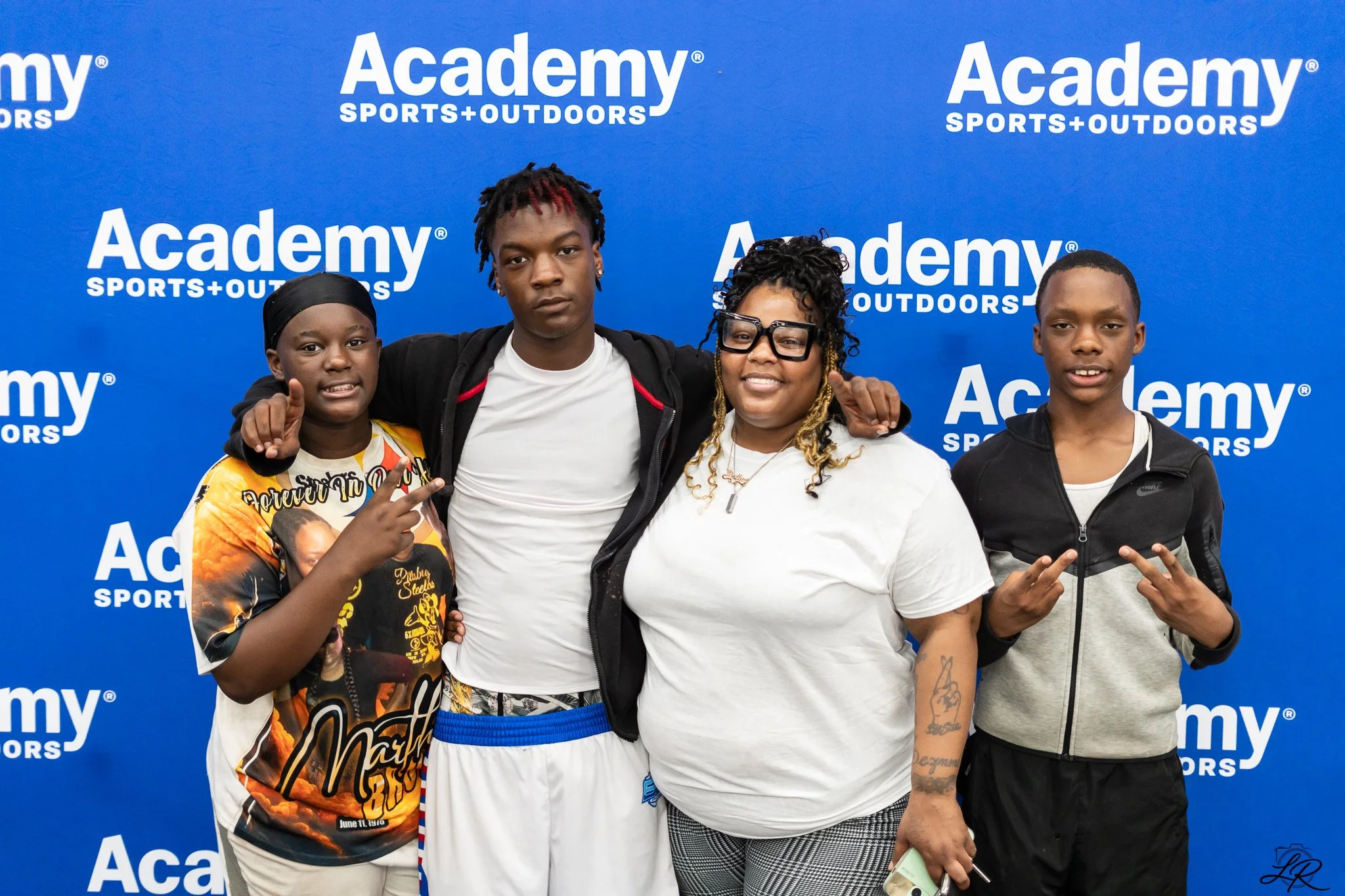 Four young people standing together in front of a blue background with the logo 'Academy Sports + Outdoors.' They are posing for a group photo, smiling, and making hand gestures.