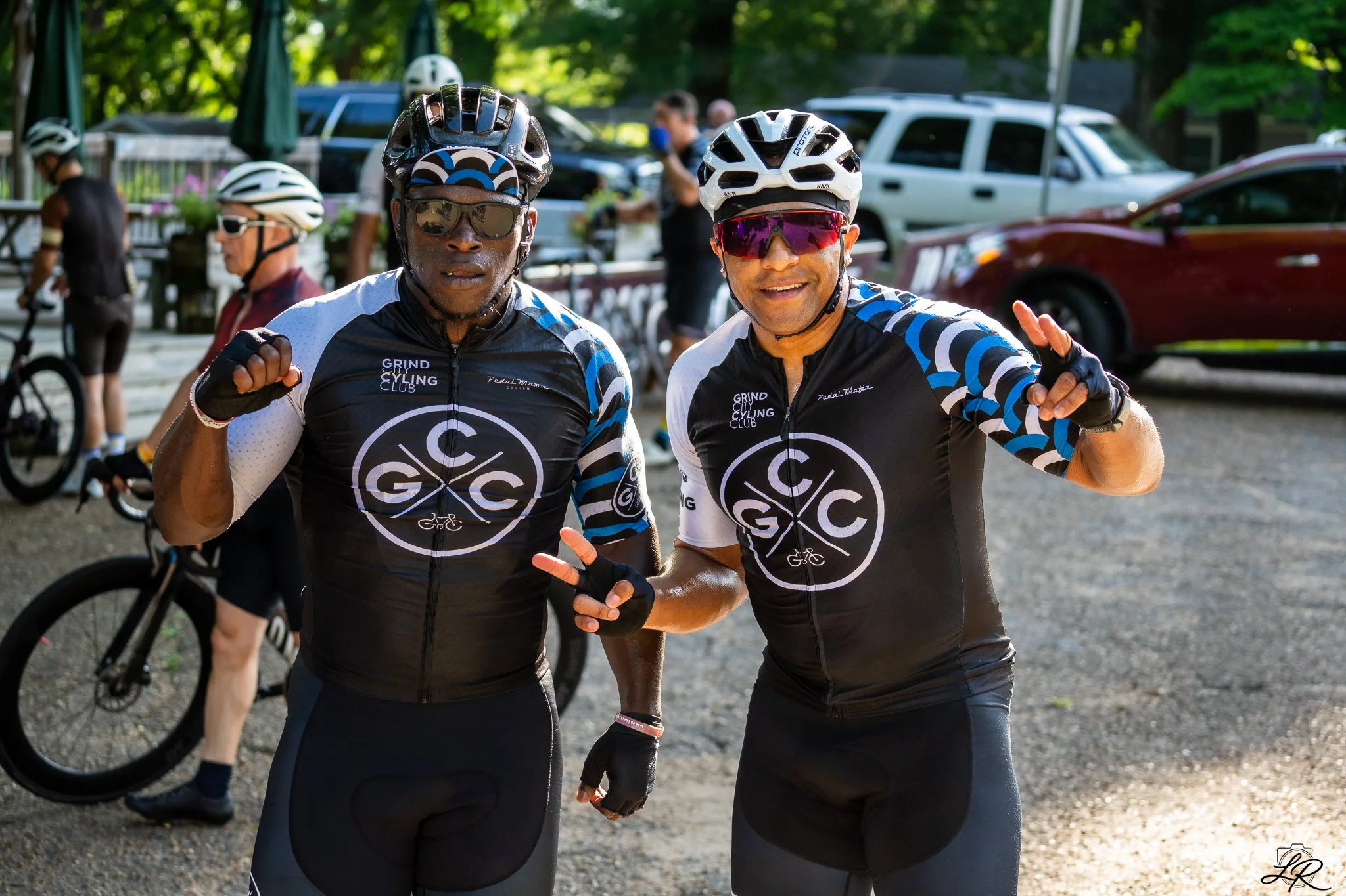Two male cyclists wearing matching black and white cycling jerseys with blue details and helmets, posing with fists near their shoulders outdoors with cars and trees in the background.