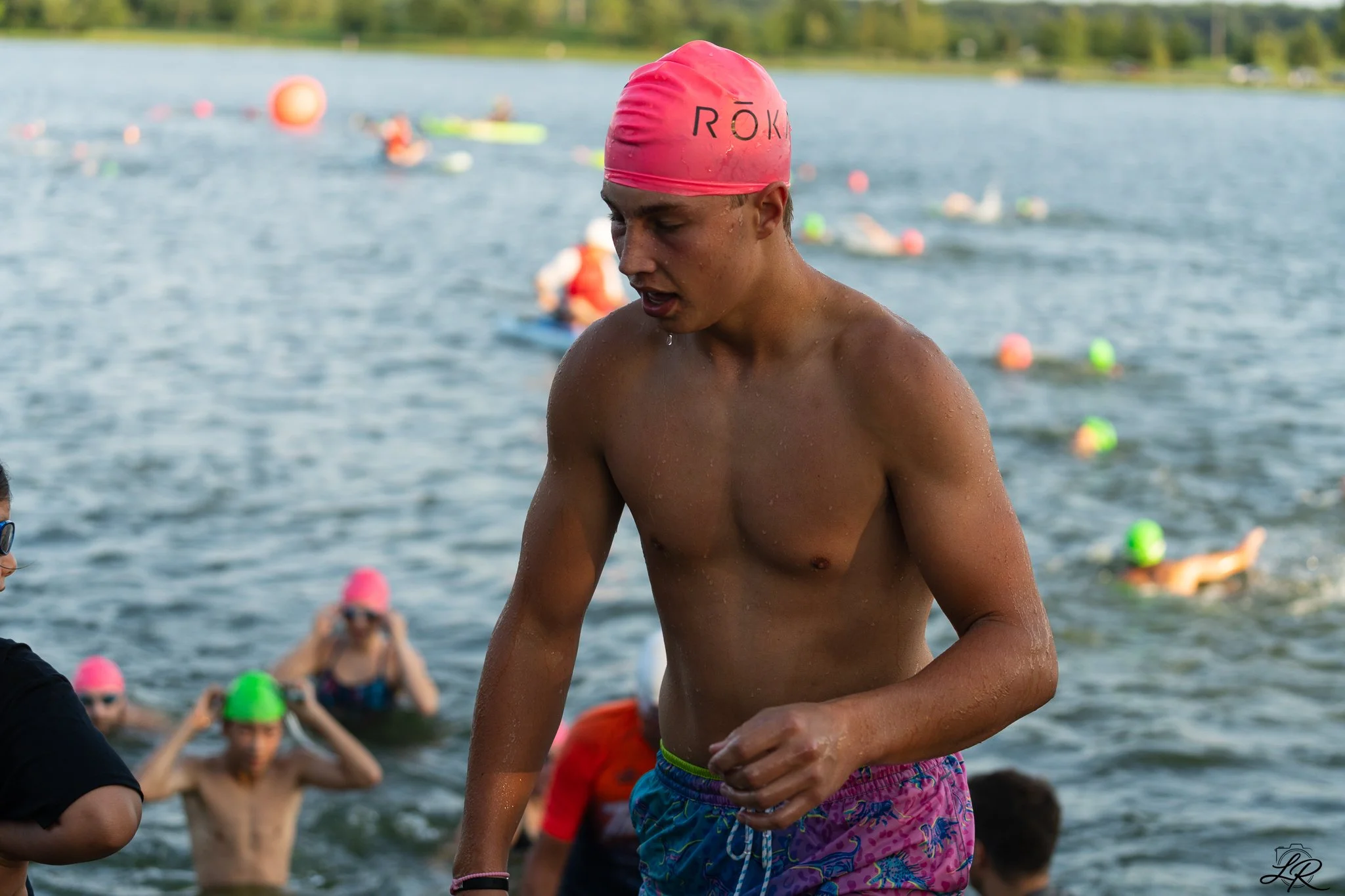 Young male swimmer wearing pink swim cap and colorful swim trunks emerging from a lake, with several other swimmers in the background.