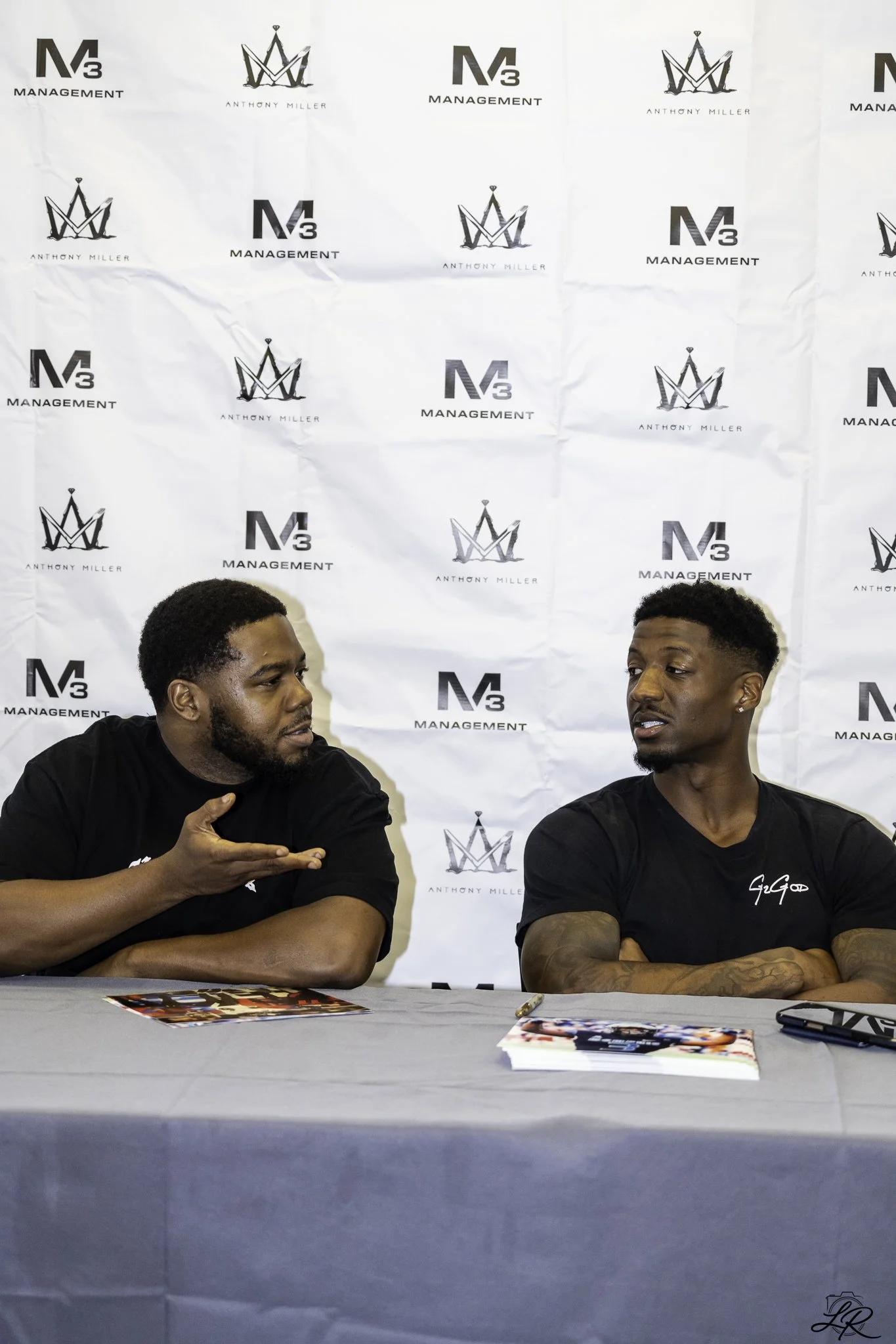 Two men sitting at a table engaged in conversation during a public event or signing. The backdrop features alternating logos of M3 Management and Anthony Miller, along with a crown symbol. On the table, there are promotional materials including a mag