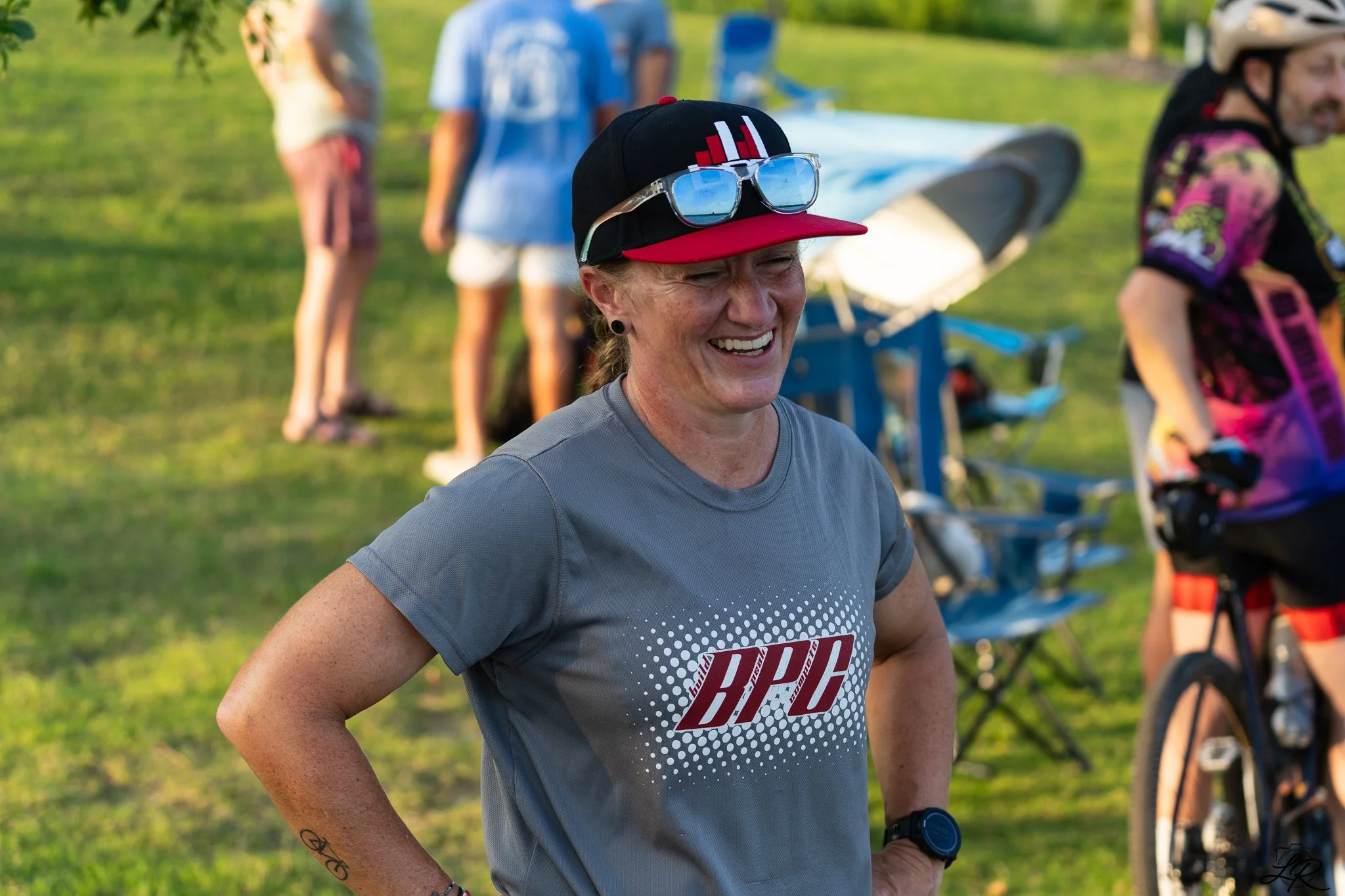 Smiling woman wearing a gray athletic shirt, a black and red cap with sunglasses on the bill, participating in an outdoor running event, with other people and bicycles in the background.