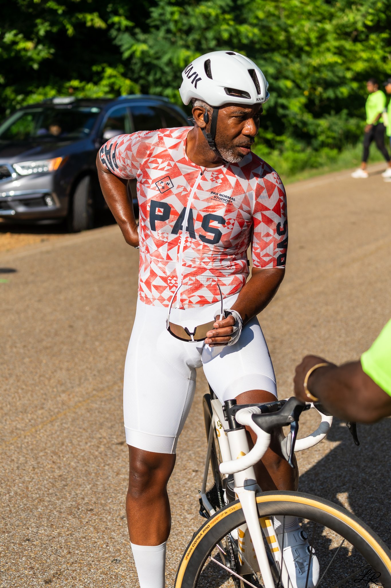 A man in a red and white geometric patterned cycling jersey and white cycling shorts, wearing a white helmet, stands next to a white bicycle with yellow tires, holding sunglasses in his left hand. Behind him is a car and a green, leafy background.