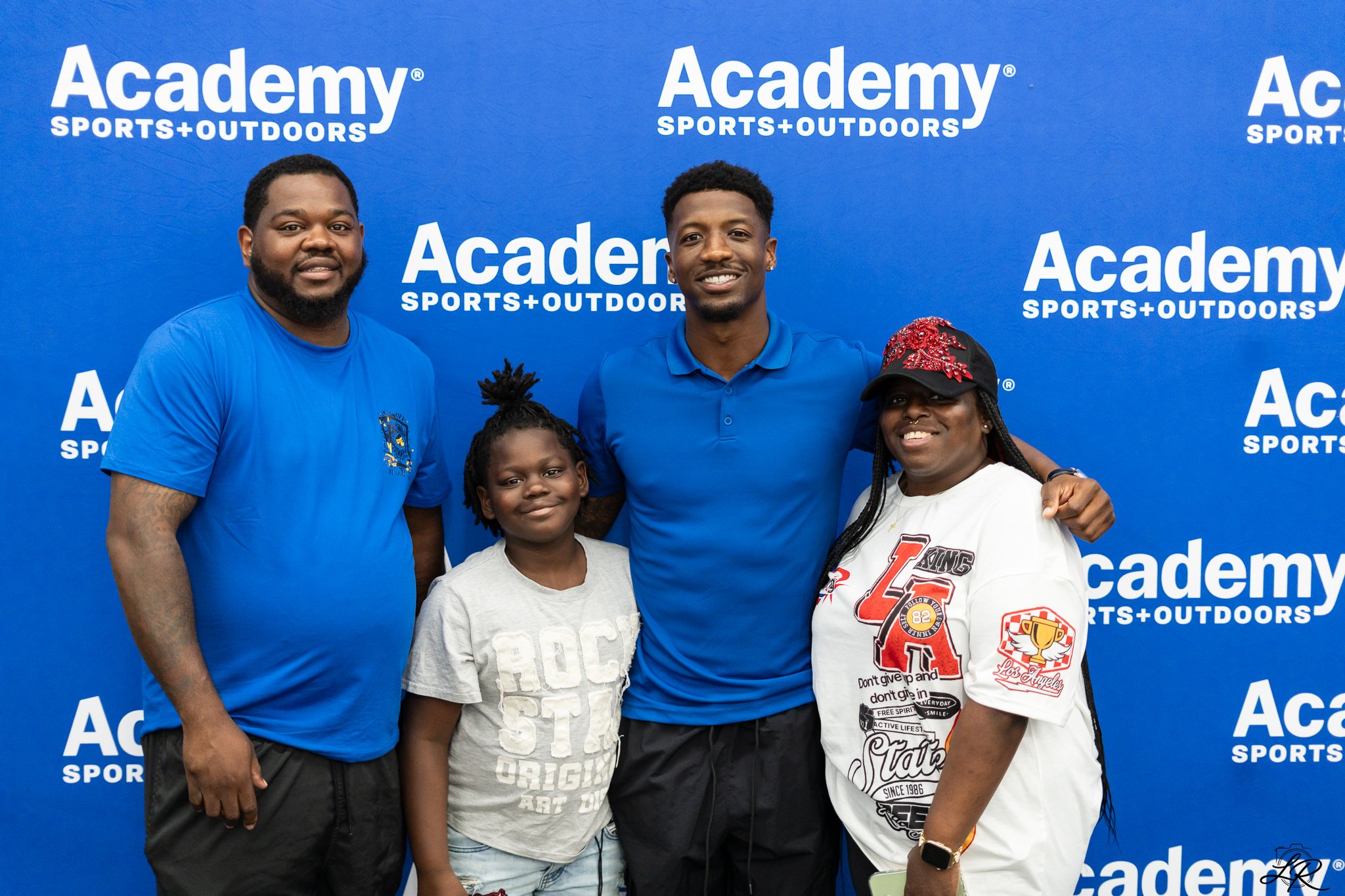 Group of four people posing together in front of a blue backdrop with white text that says 'Academy Sports + Outdoors'.