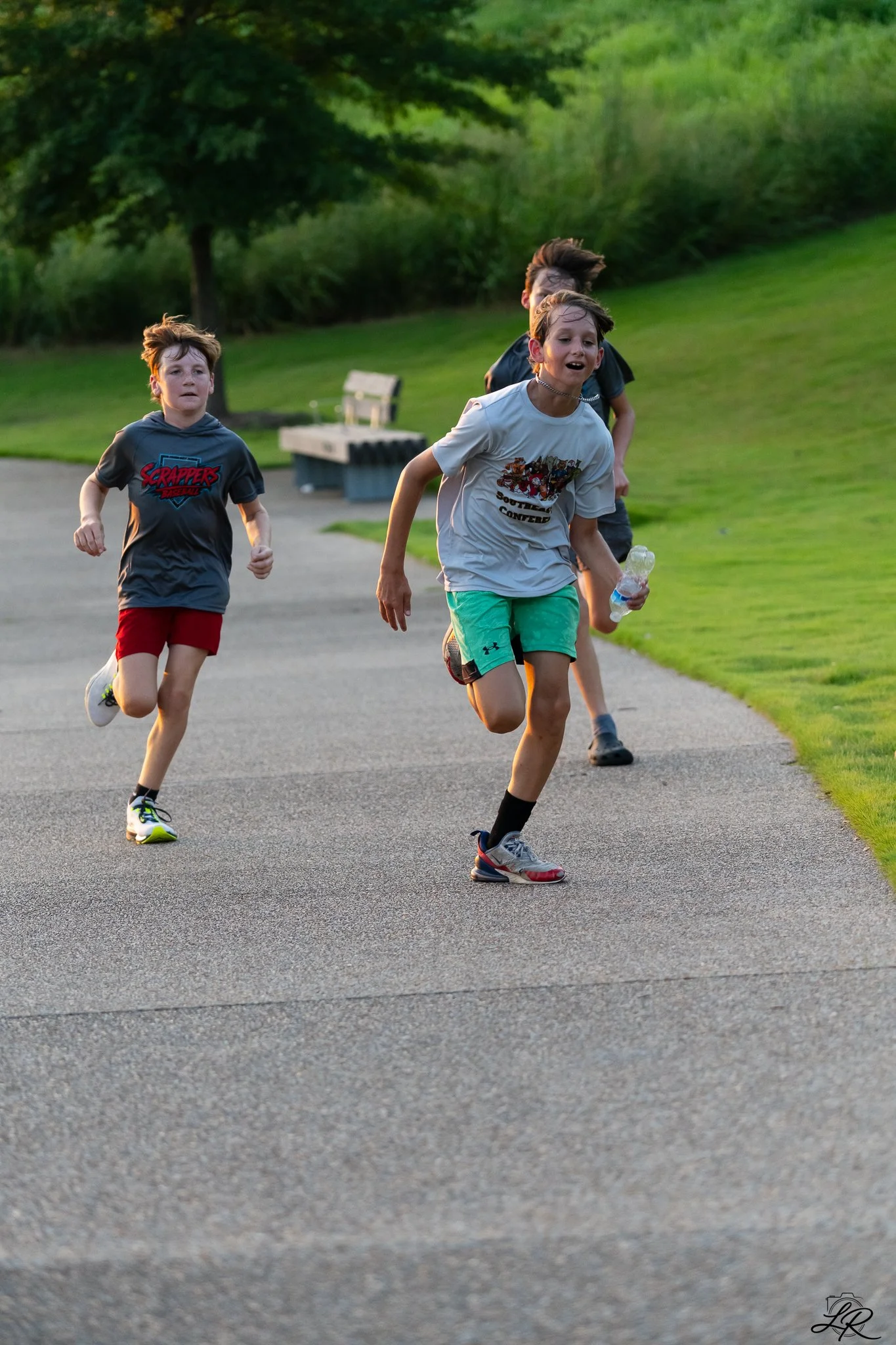 Children running outdoors on a paved path in a park with lush green grass and trees in the background, late afternoon or early evening light.