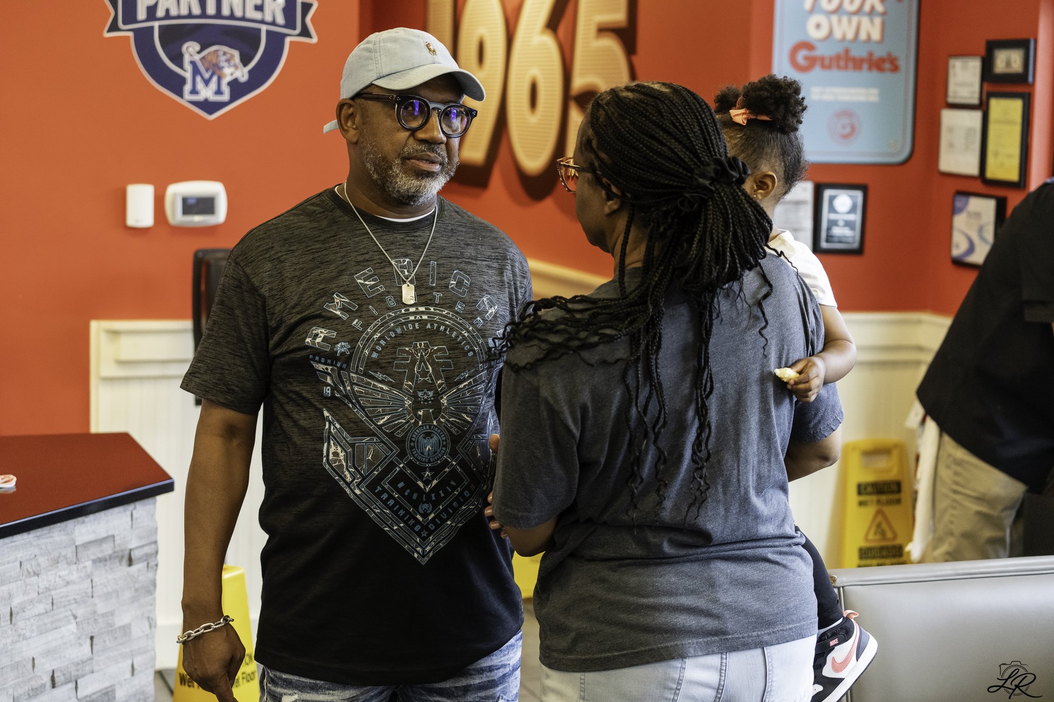 An older man wearing glasses, a light gray cap, and a graphic T-shirt is engaging in conversation with a woman with braided hair and glasses in a fast-food restaurant. The background features red walls with signs, including a large sign that reads 'P