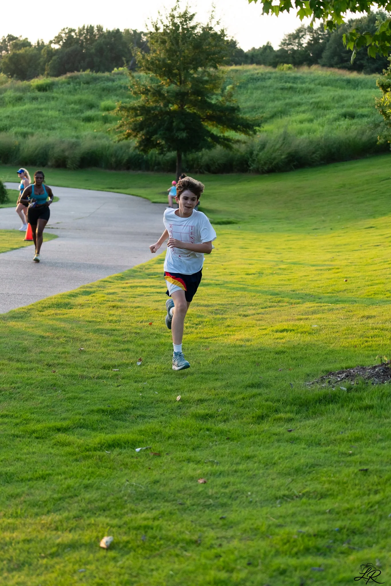 Children running on a grassy park trail in the late afternoon, greenery and trees in the background.