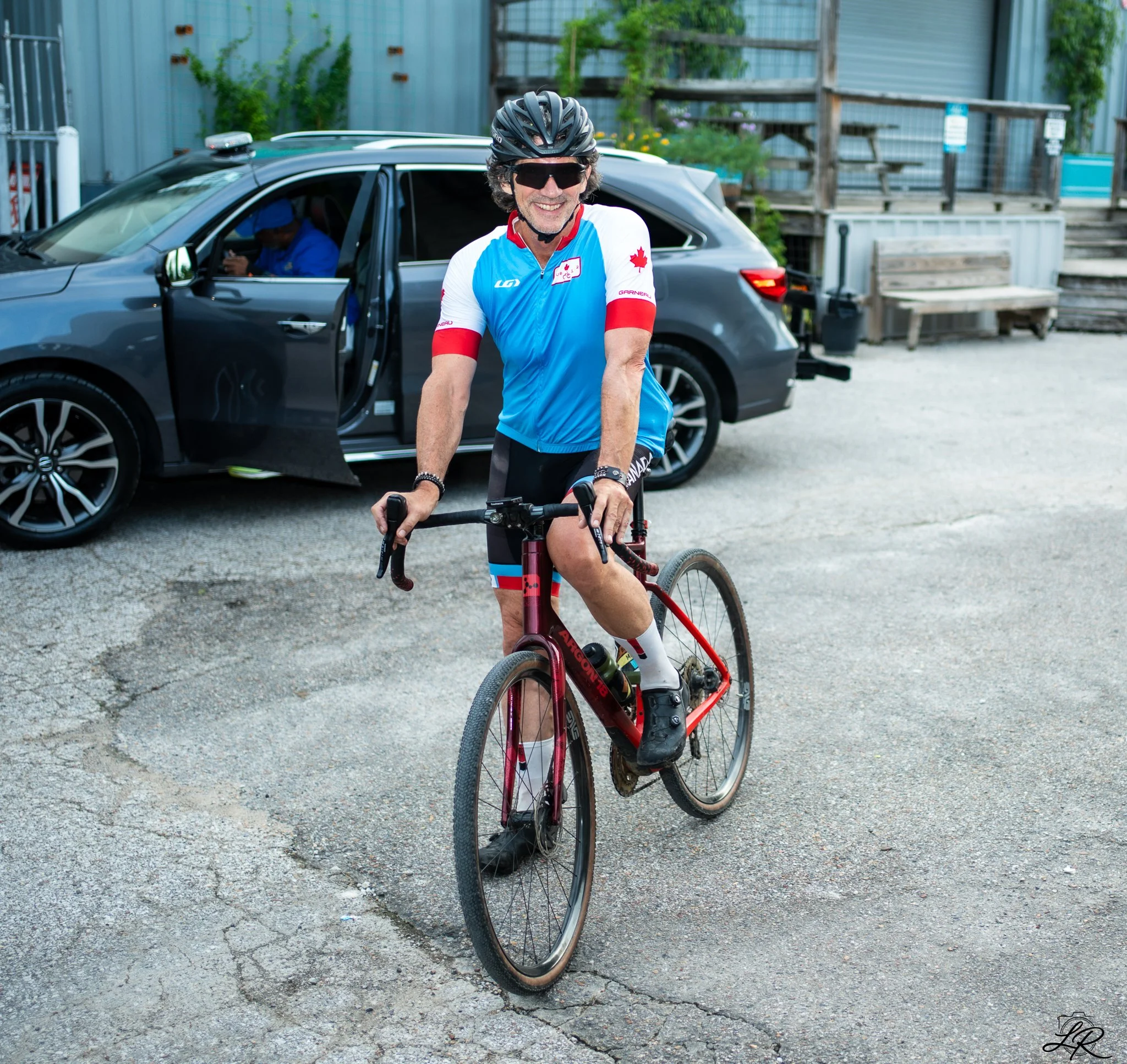 A man dressed in cycling gear, including a Canadian flag jersey, helmet, and sunglasses, standing next to a red mountain bike on a gravel surface. A black SUV with an open door is in the background, with a person sitting inside using a smartphone. Th