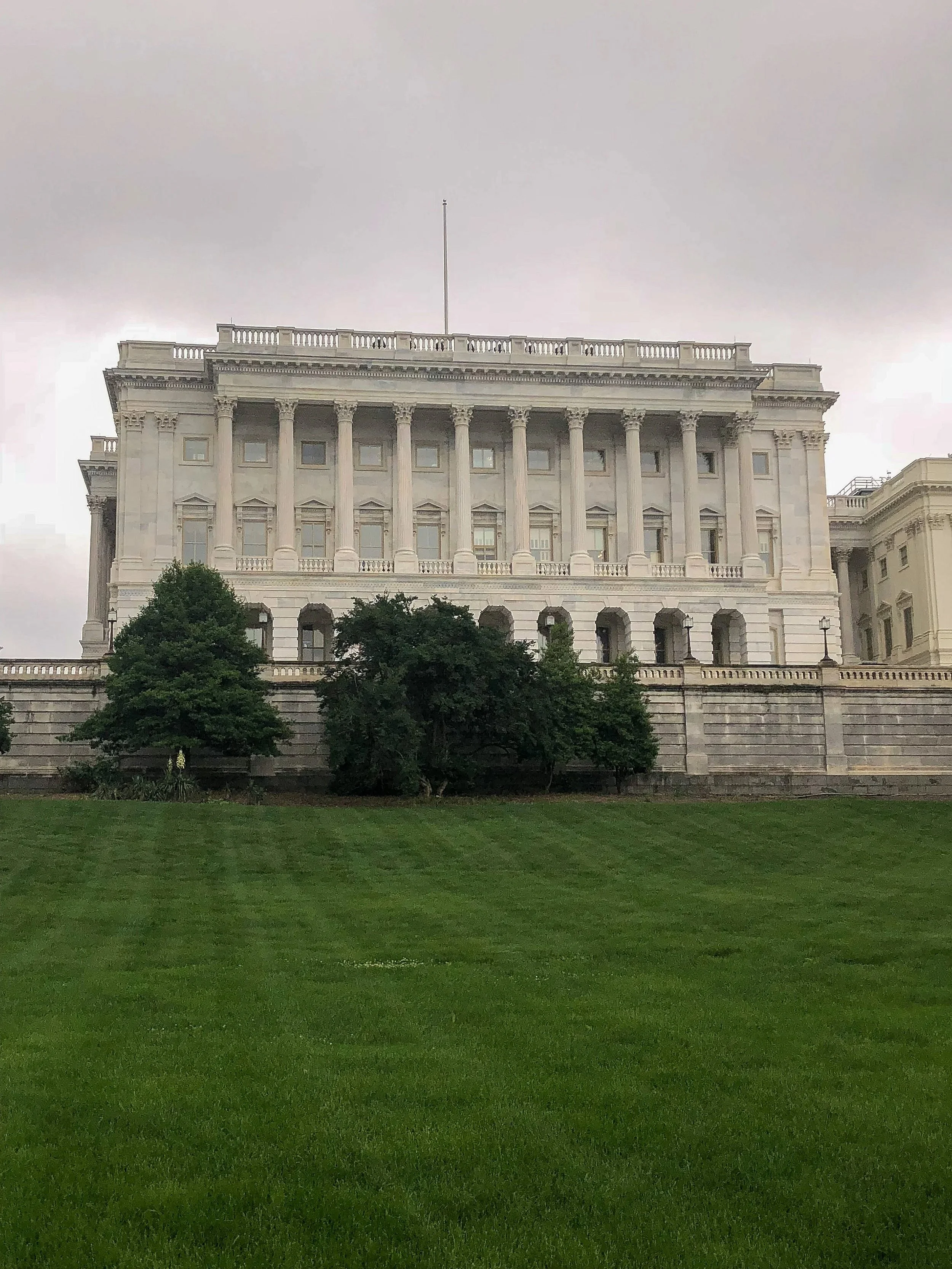 The United States Capitol building seen from the front, with a well-maintained green lawn in the foreground and trees partially obscuring the lower part of the building. The sky is cloudy.