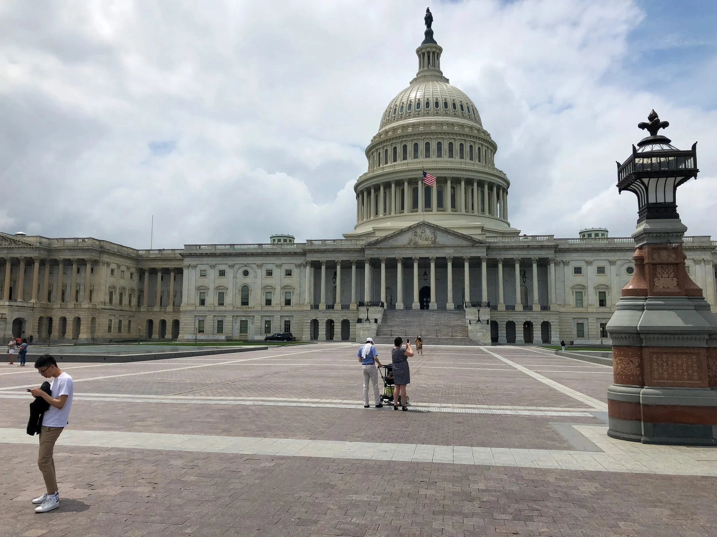 The United States Capitol building with a large dome, American flag, and several people in front, some taking photos, under a cloudy sky.