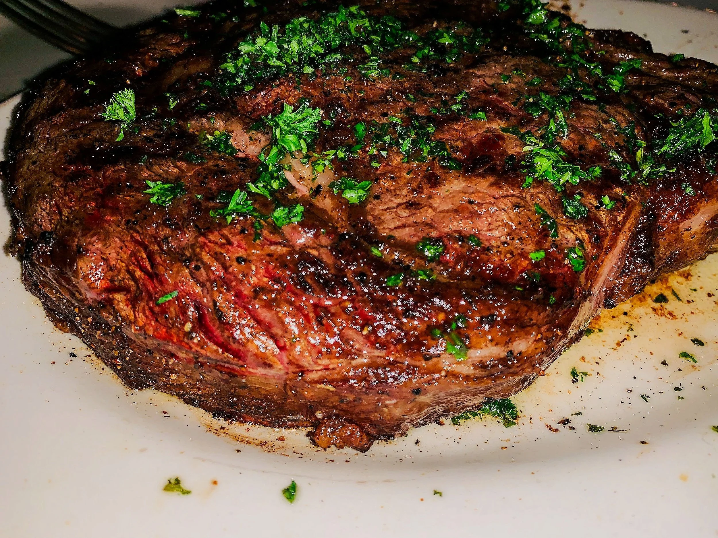 A cooked ribeye steak garnished with chopped parsley, placed on a white plate.
