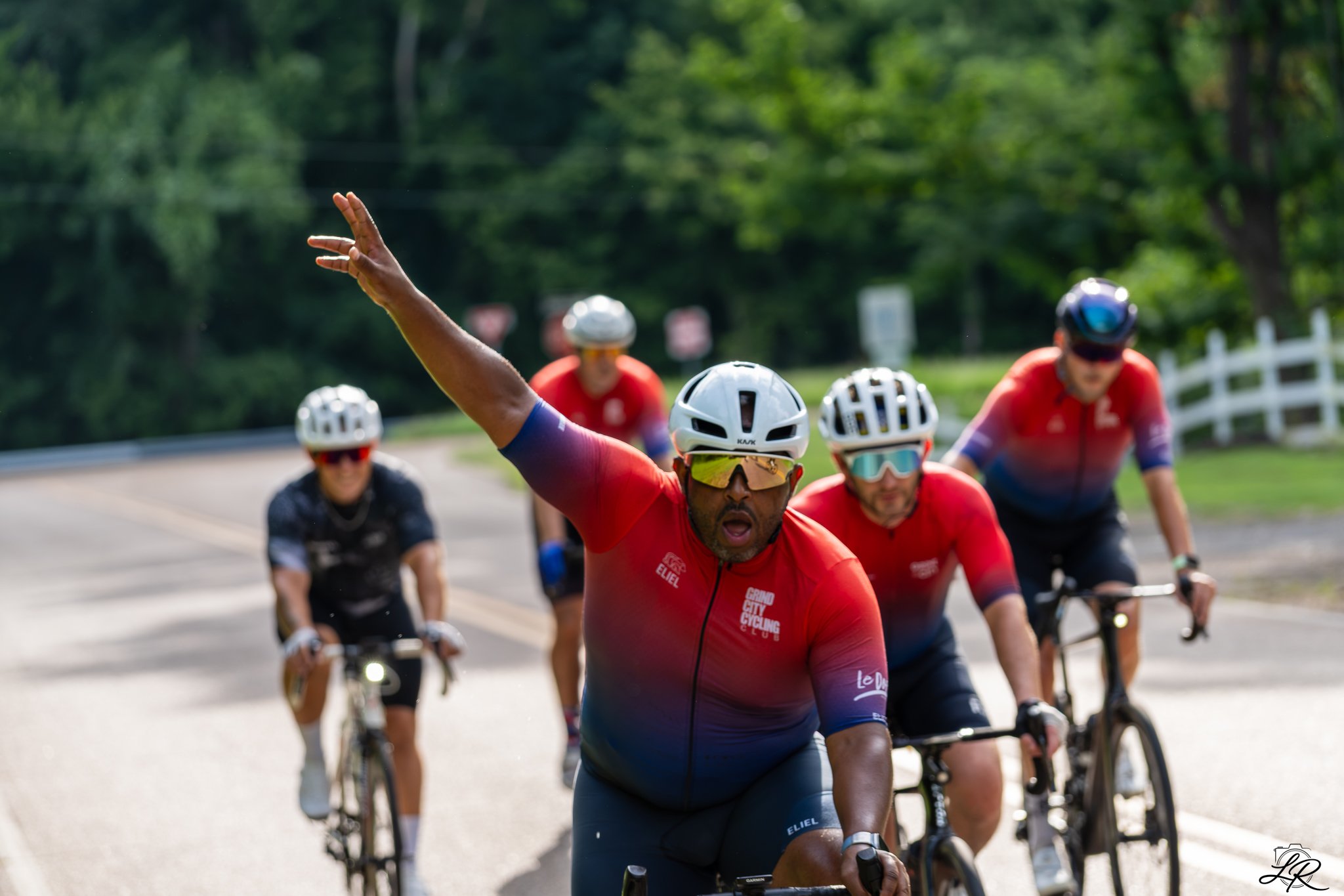 A group of male cyclists in red and black jerseys riding on a road through a green, wooded area. One cyclist in front is raising his right arm and appears to be leading or celebrating.