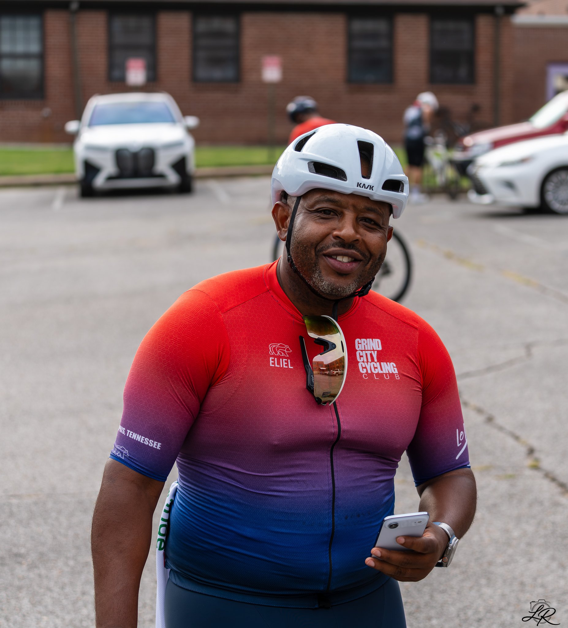 A man wearing a red and blue cycling jersey and a white helmet is looking at his phone while standing outside. In the background, there are parked cars and a brick building.