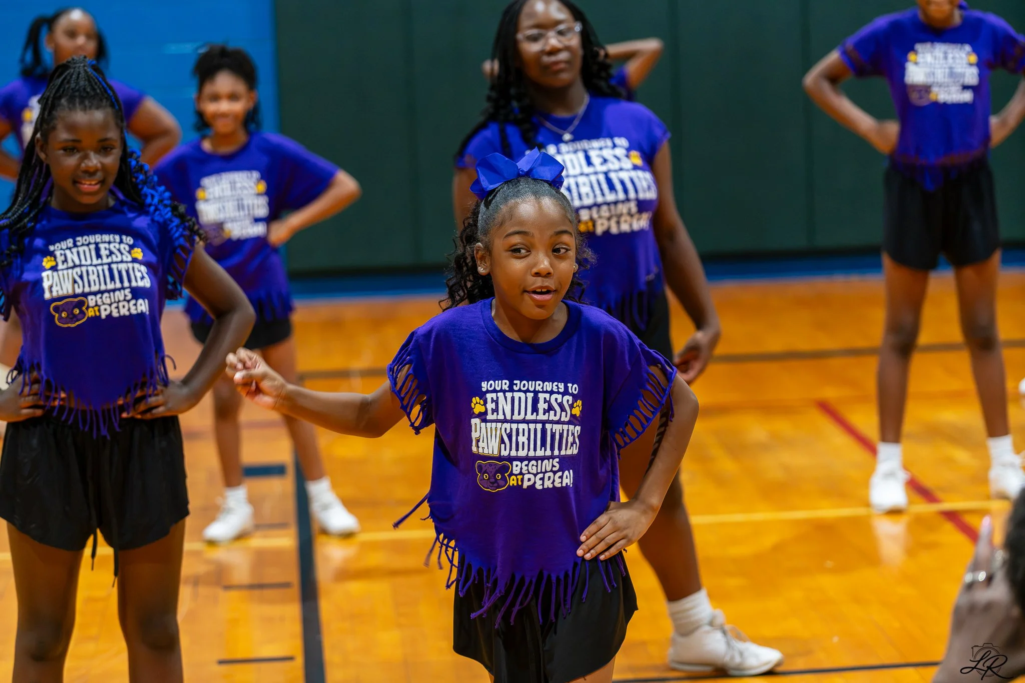 A group of young girls participating in a cheerleading or dance practice in a gymnasium. They wear matching purple shirts with white text and black skirts, with one girl in the foreground with a blue bow in her hair. The background is blurred but sho