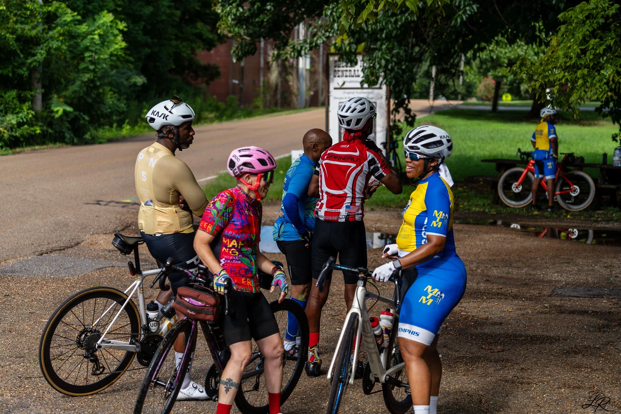 Group of cyclists, including men and women, gathered and talking outdoors with trees and a sign in the background