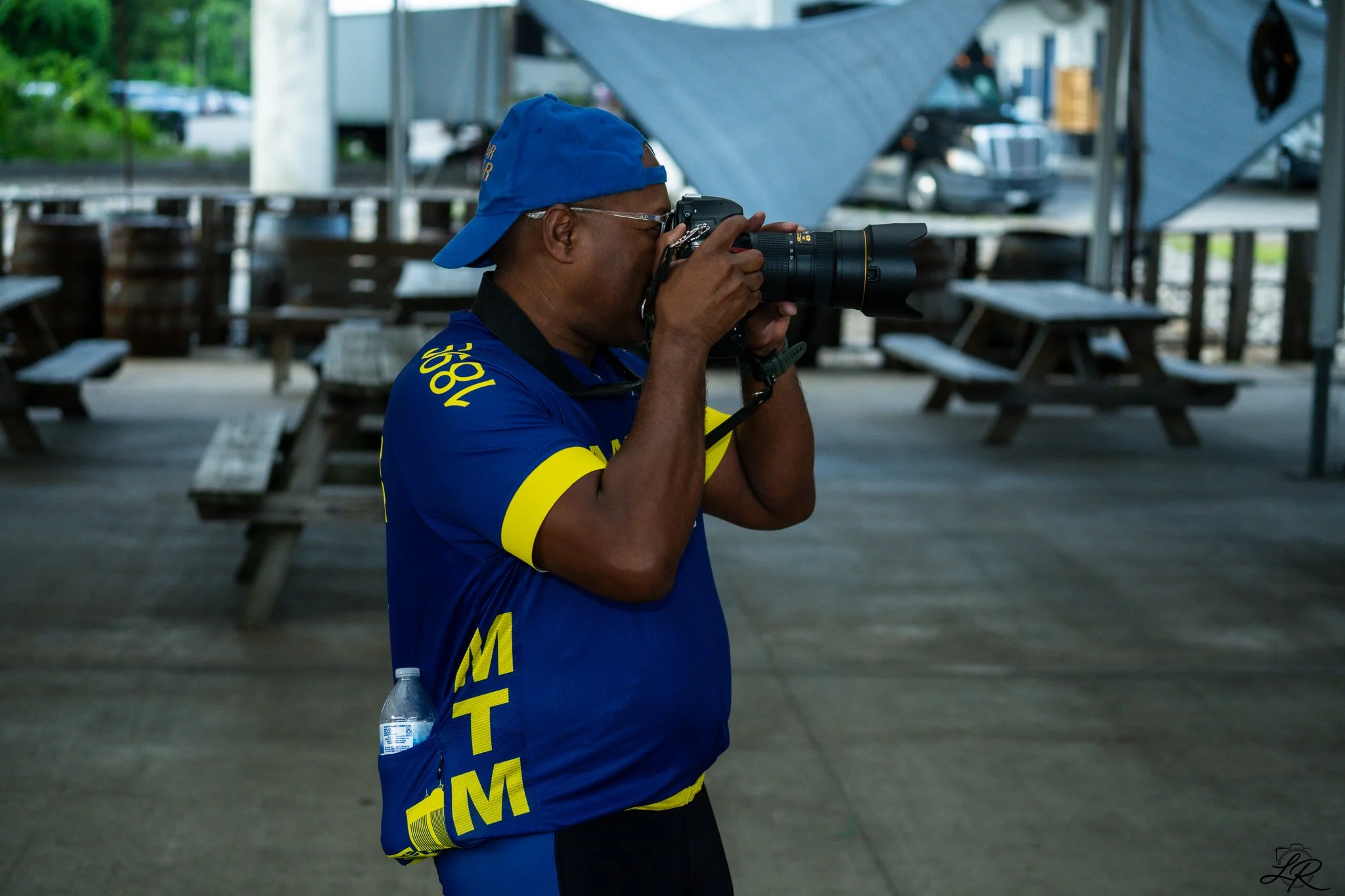 A man wearing a blue cap and a blue shirt with yellow lettering is taking a photograph with a camera equipped with a zoom lens. He appears to be outdoors, standing next to picnic tables and a parking lot in the background.