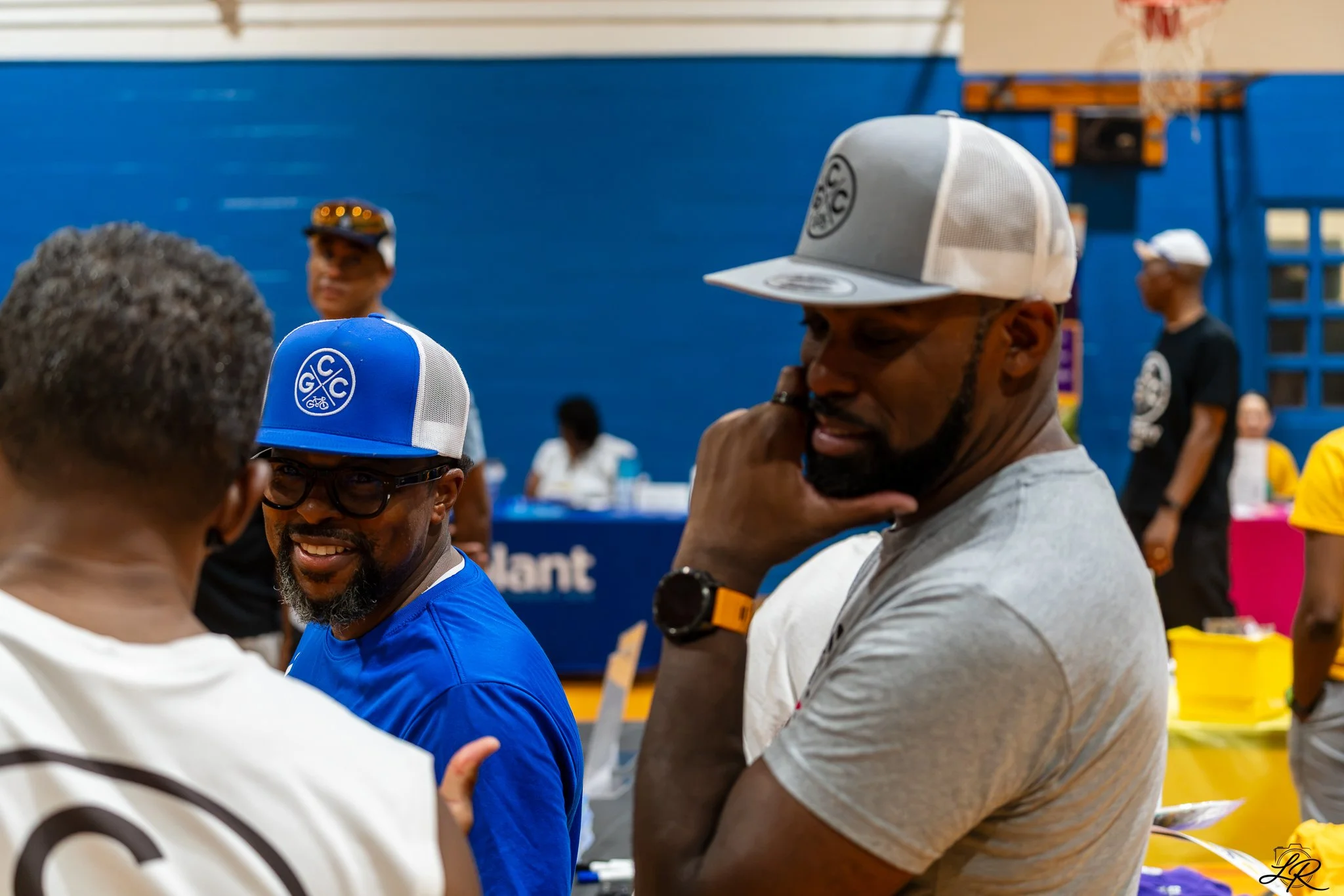 A group of people at an indoor event, some wearing colorful caps and casual clothing, standing and engaging in conversations, with a blue wall and a basketball hoop in the background.