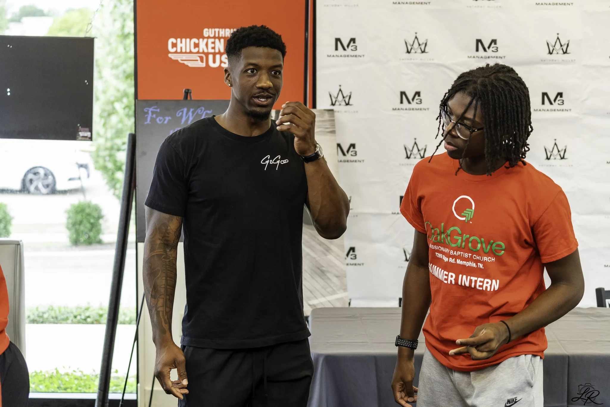 Two young men standing and talking indoors at a promotional or community event, with a banner in the background displaying multiple logos and text.