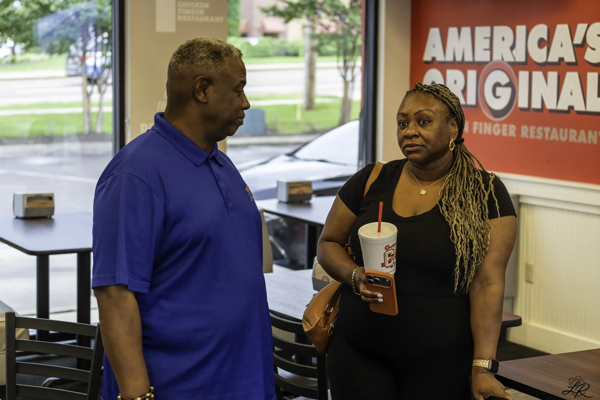 A man in a blue shirt and a woman with braided hair and a black shirt stand inside a restaurant, engaging in conversation. The woman holds a drink with a straw and a cell phone in her hand. A red sign behind them reads 'America's Original.'