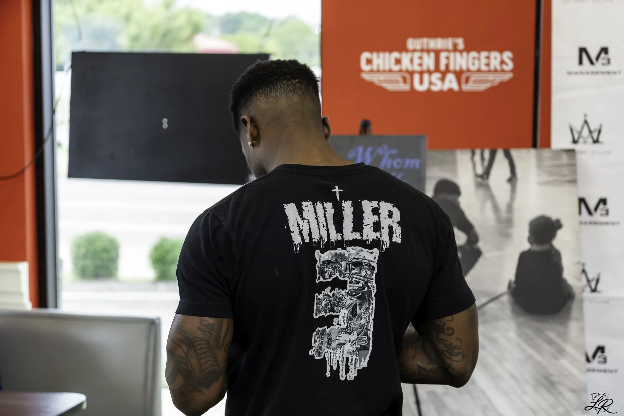 Person with tattoos wearing a black t-shirt with a graphic design on the back, standing indoors near a window, in front of printed displays including one with a red banner that reads 'Butcher's Chicken Fingers USA'.