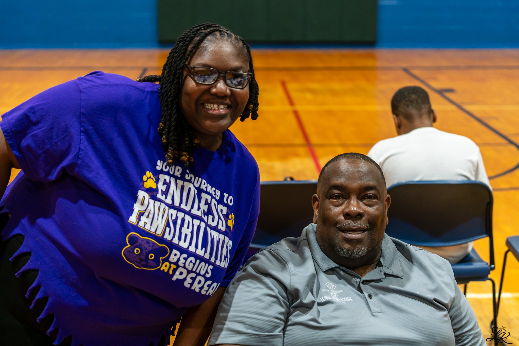Smiling woman wearing glasses and a blue shirt with text, sitting next to a man in a gray shirt inside a gymnasium.