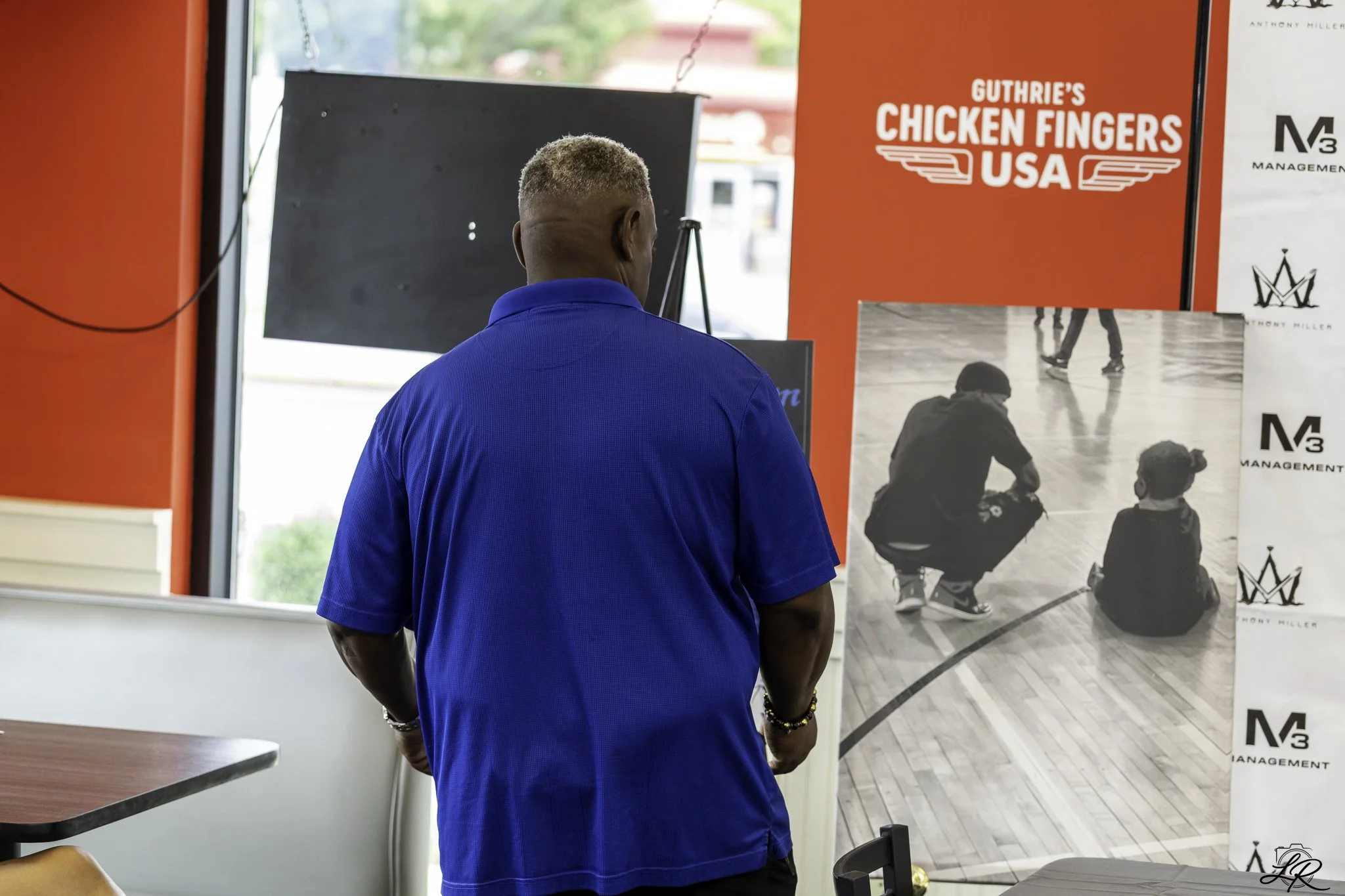 A man with short blonde hair wearing a blue polo shirt standing indoors near a large window. Behind him, a red sign reads "Guthrie's Chicken Fingers USA." To his right, there are black and white photographs of children playing hockey, along with the 