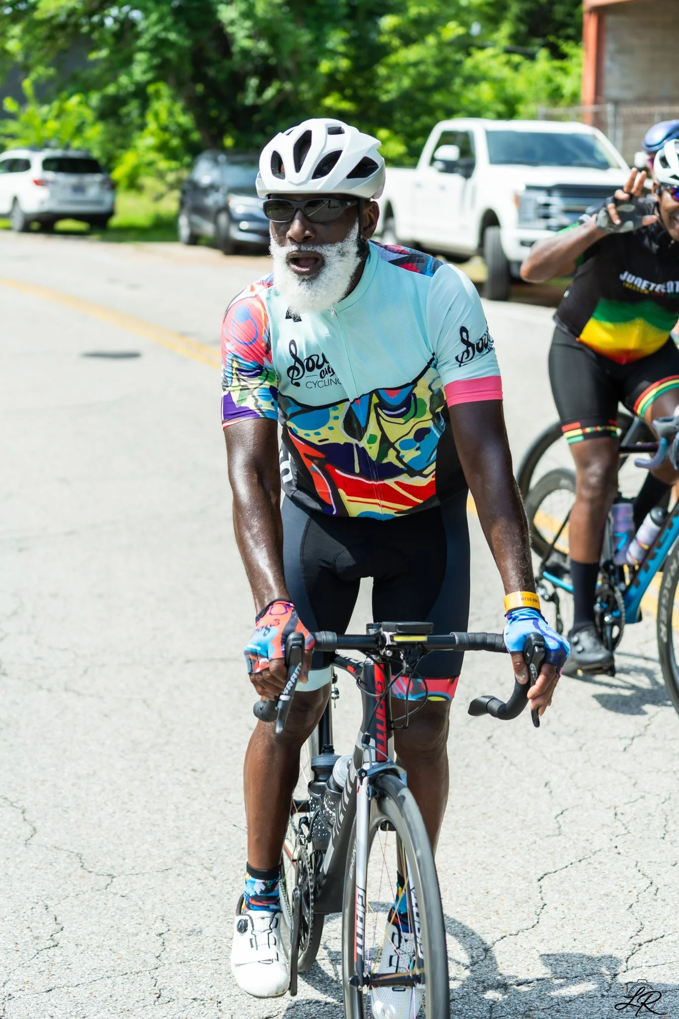 A man in colorful cycling attire, wearing a white helmet and sunglasses, riding a bicycle on a cracked asphalt road with parked cars and green trees in the background.