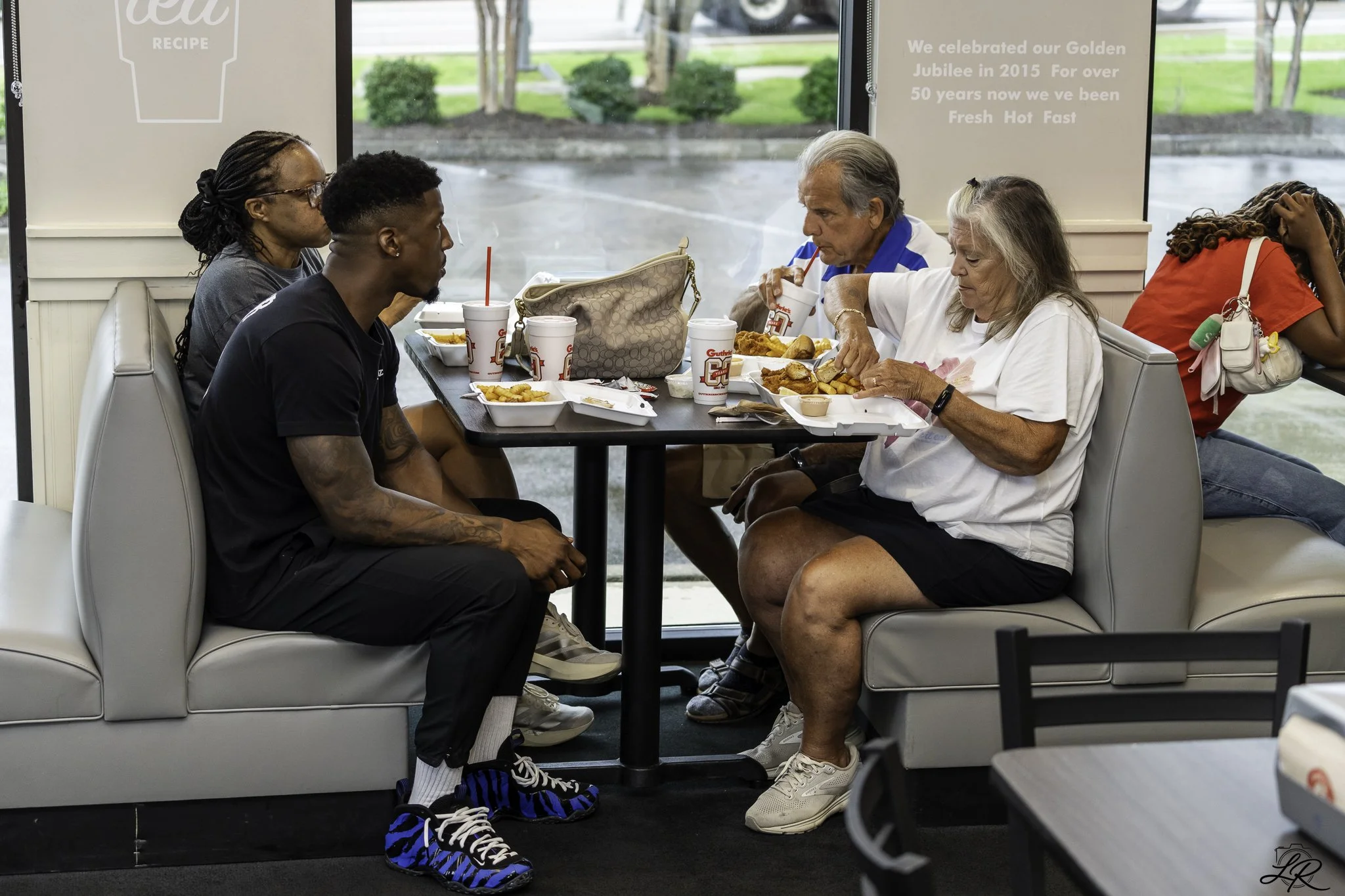 Six people sitting at a table in a fast-food restaurant, eating meals. The group includes women with diverse hair styles and ages, with one woman with gray hair and a man with gray hair, all enjoying their food. The restaurant has large windows and p