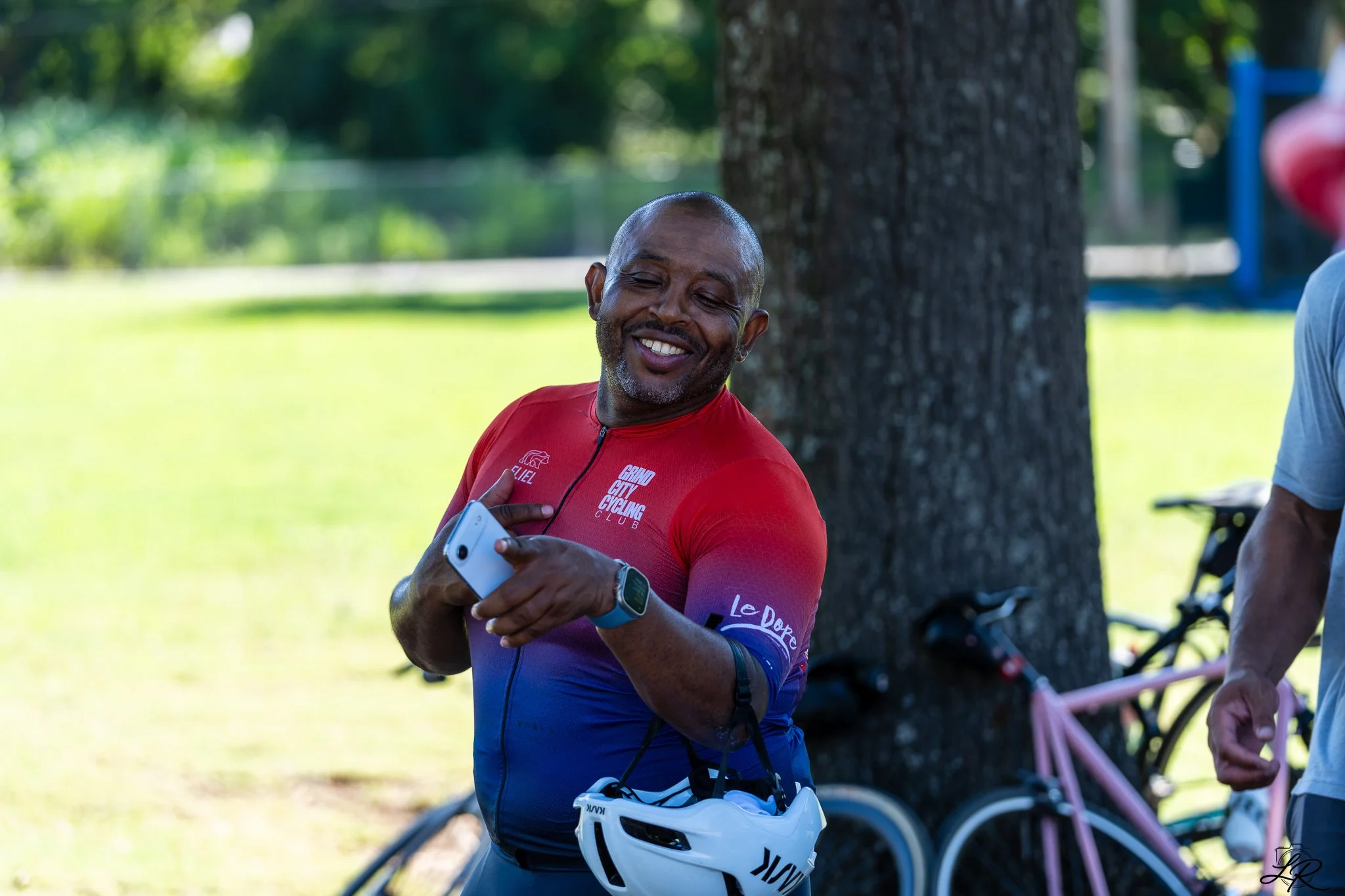 A smiling man in a red cycling jersey holding a smartphone, standing outdoors near a tree, with a white helmet hanging from his hand. There are cycling shoes, a bicycle, and other equipment in the background.
