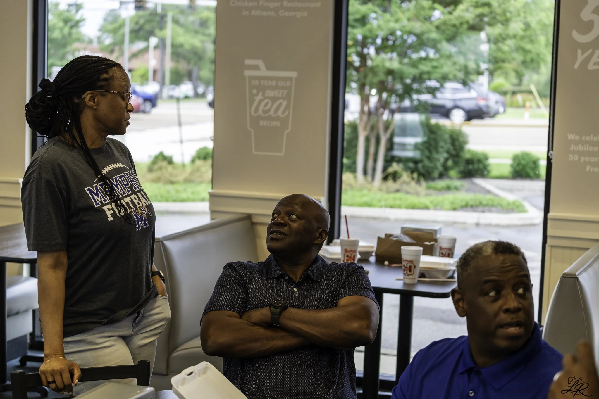 Four people inside a restaurant near a window, with trees and parked cars visible outside. One woman is standing wearing glasses and a Memphis football t-shirt, looking at a man sitting across from her who is looking up at her. Two other men are seat