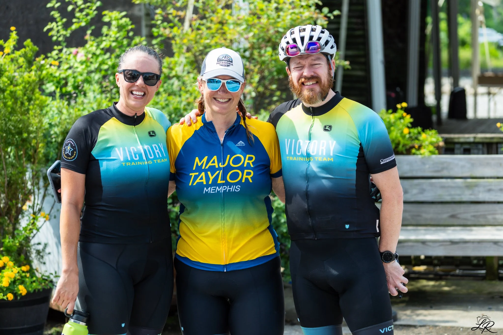 Three people in cycling gear standing outdoors smiling for the camera. The woman in the middle is wearing a blue and yellow cycling jersey that reads 'Major Taylor Memphis', while the two people on either side are wearing black and yellow jerseys tha