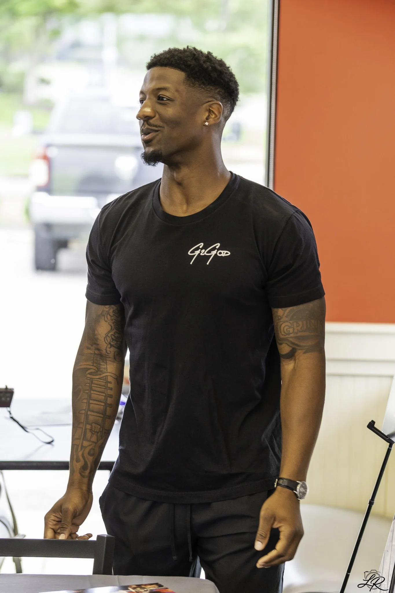 A young man with short curly hair and tattoos on his arms, wearing a black t-shirt with the words 'Go Go' on it, is standing indoors near a window with blurred cars outside. He appears to be smiling or talking.