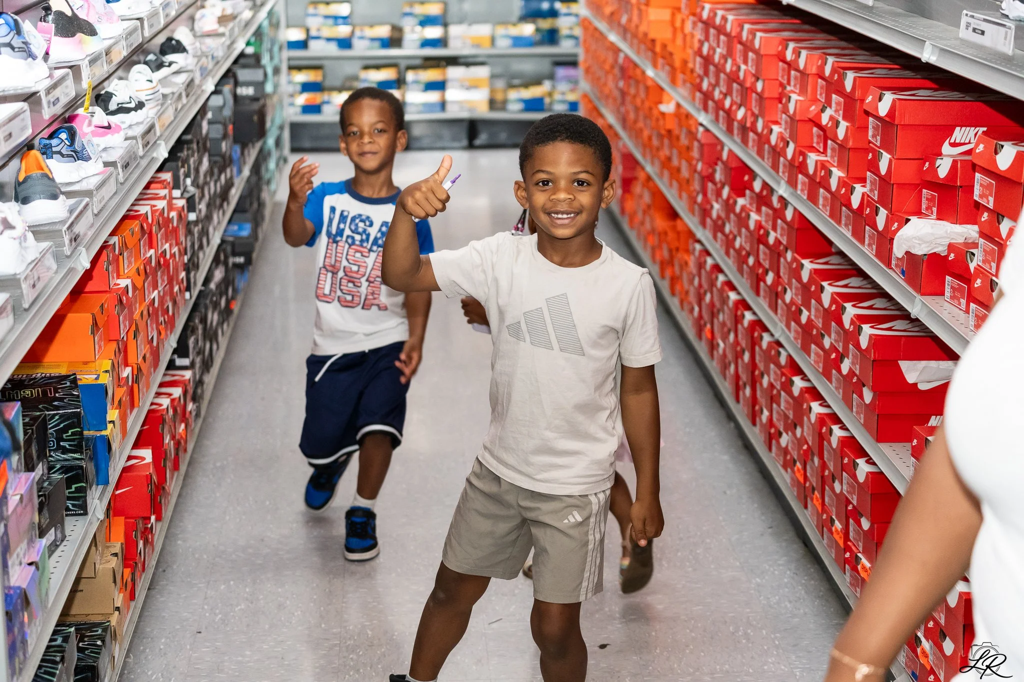 Two young boys shopping for sneakers in a store aisle lined with Nike shoeboxes. One boy giving a thumbs-up, smiling, while the other boy runs behind.