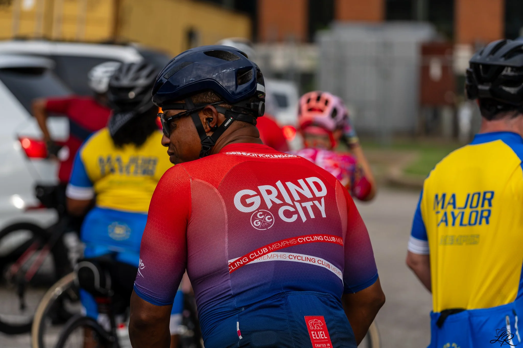 Close-up of a cyclist wearing a red and purple jersey with text 'GRAND CITY' and a helmet, standing among other cyclists in colorful jerseys and helmets, with parked cars and buildings in the background.