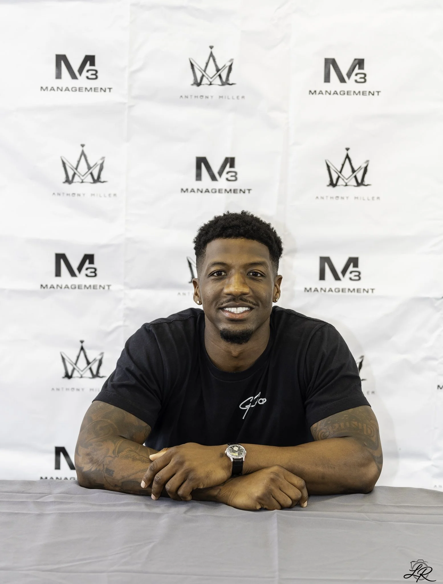 A young man with short curly hair and earrings, sitting at a table with his arms crossed. He is wearing a black t-shirt, a wristwatch, and has tattoos on his arms. Behind him is a backdrop with logos and text reading 'M3 MANAGEMENT' and 'ANTHONY MILL