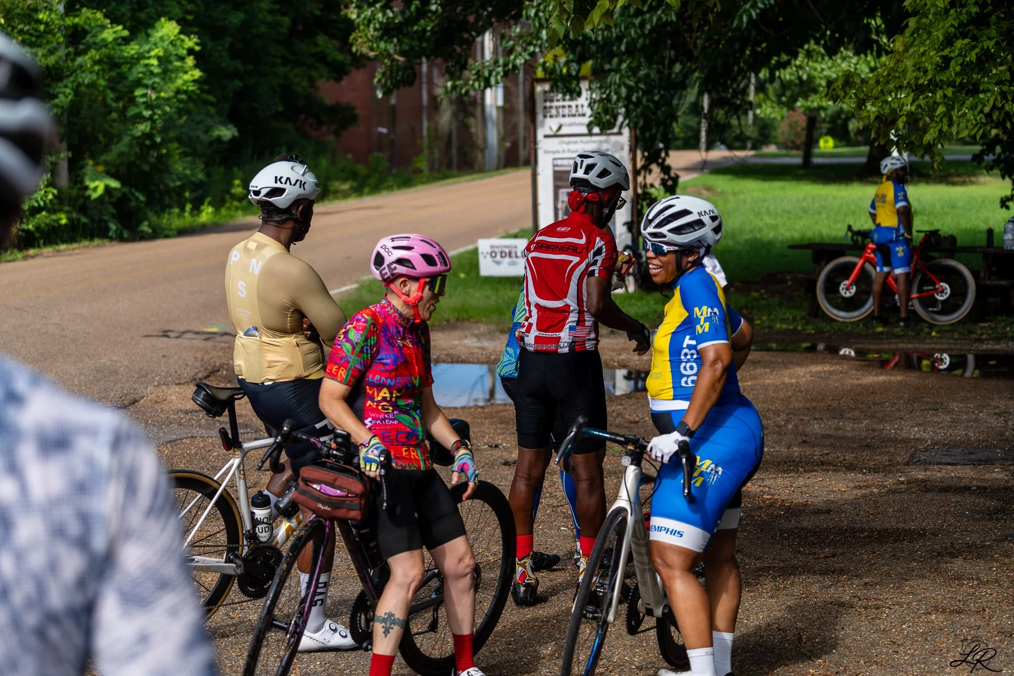 Group of cyclists in colorful jerseys and helmets talking and preparing to ride on a suburban street.