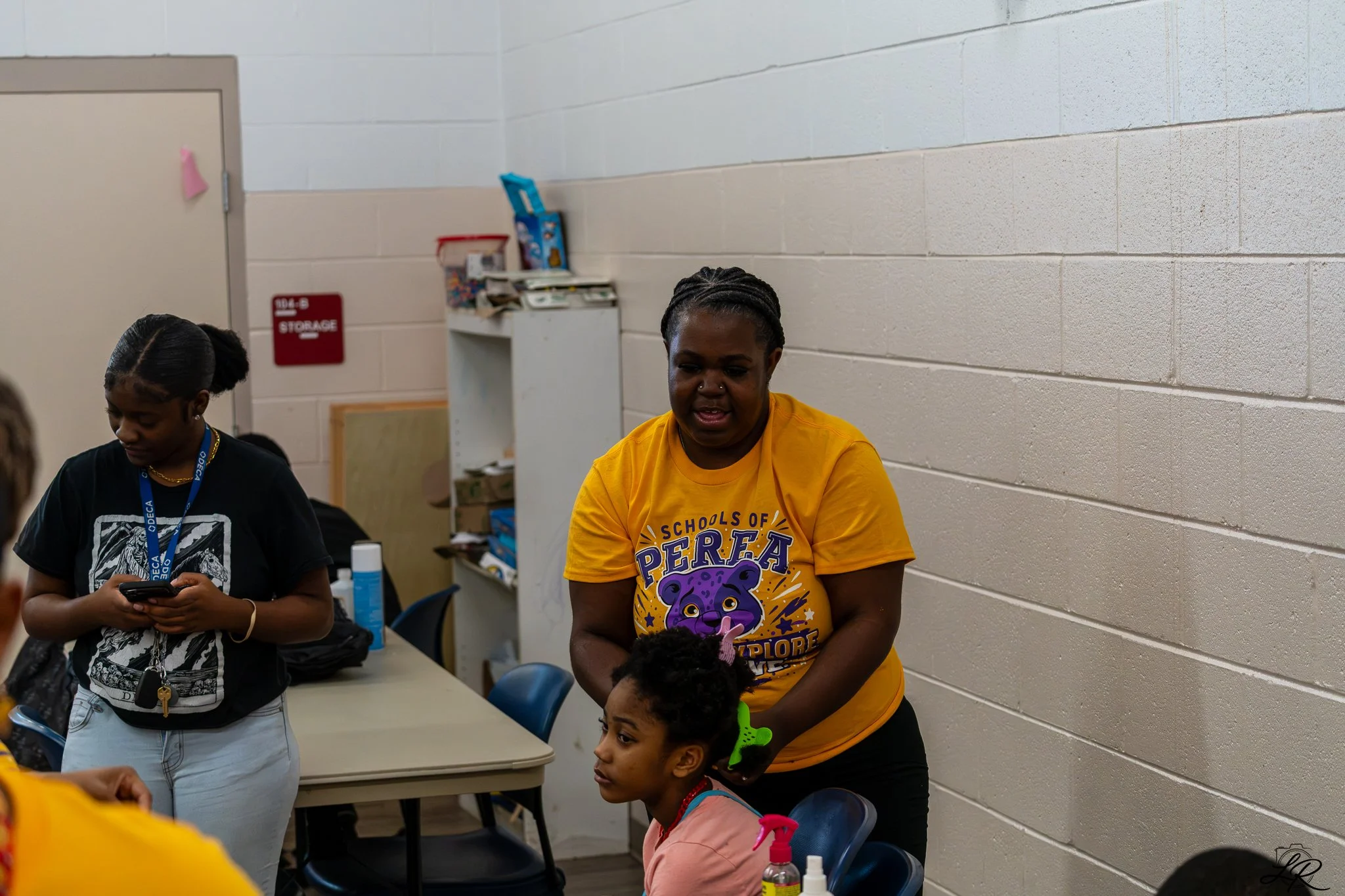 A group of children and an adult woman with dark skin in a classroom setting. The woman is wearing a yellow t-shirt with a purple bear graphic and the words "Schools of PERA". One girl is looking at her phone, while another girl with a green hair cli