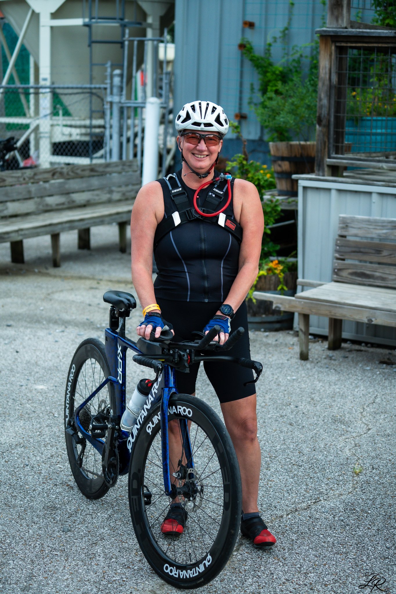 A woman smiling outdoors with a bicycle. She is wearing a white helmet, sunglasses, a black sleeveless athletic top, black shorts, and red and black cycling shoes. There are plants and wooden structures in the background.