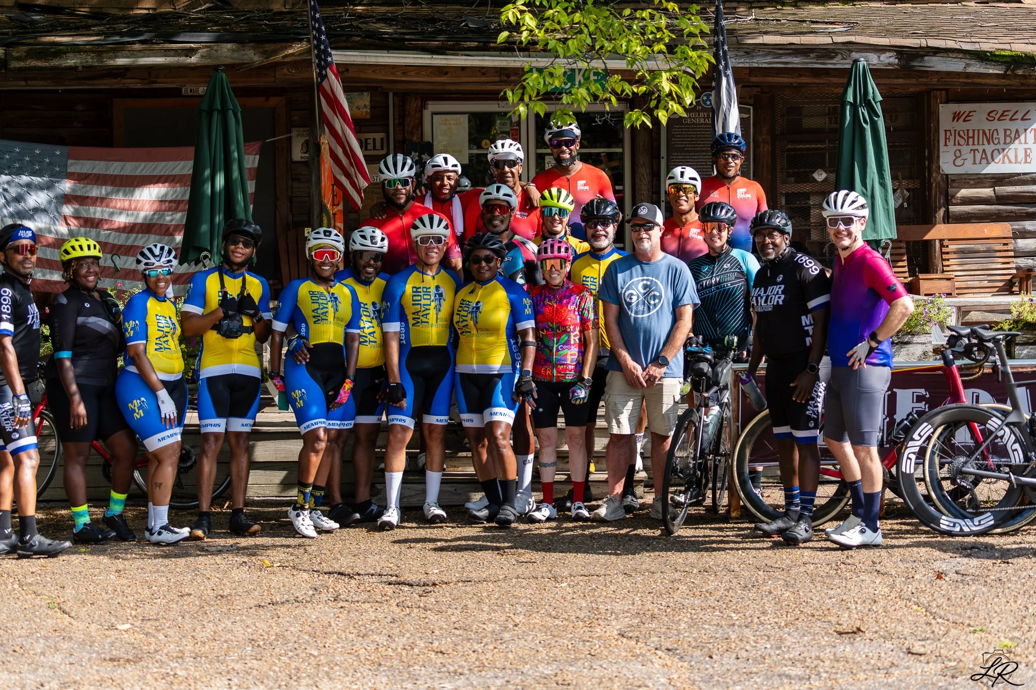 Group of cyclists and supporters posing for a photo outside a rustic building with American flags, umbrellas, and a sign that says 'We Sell Fishing Bait & Tackle'.