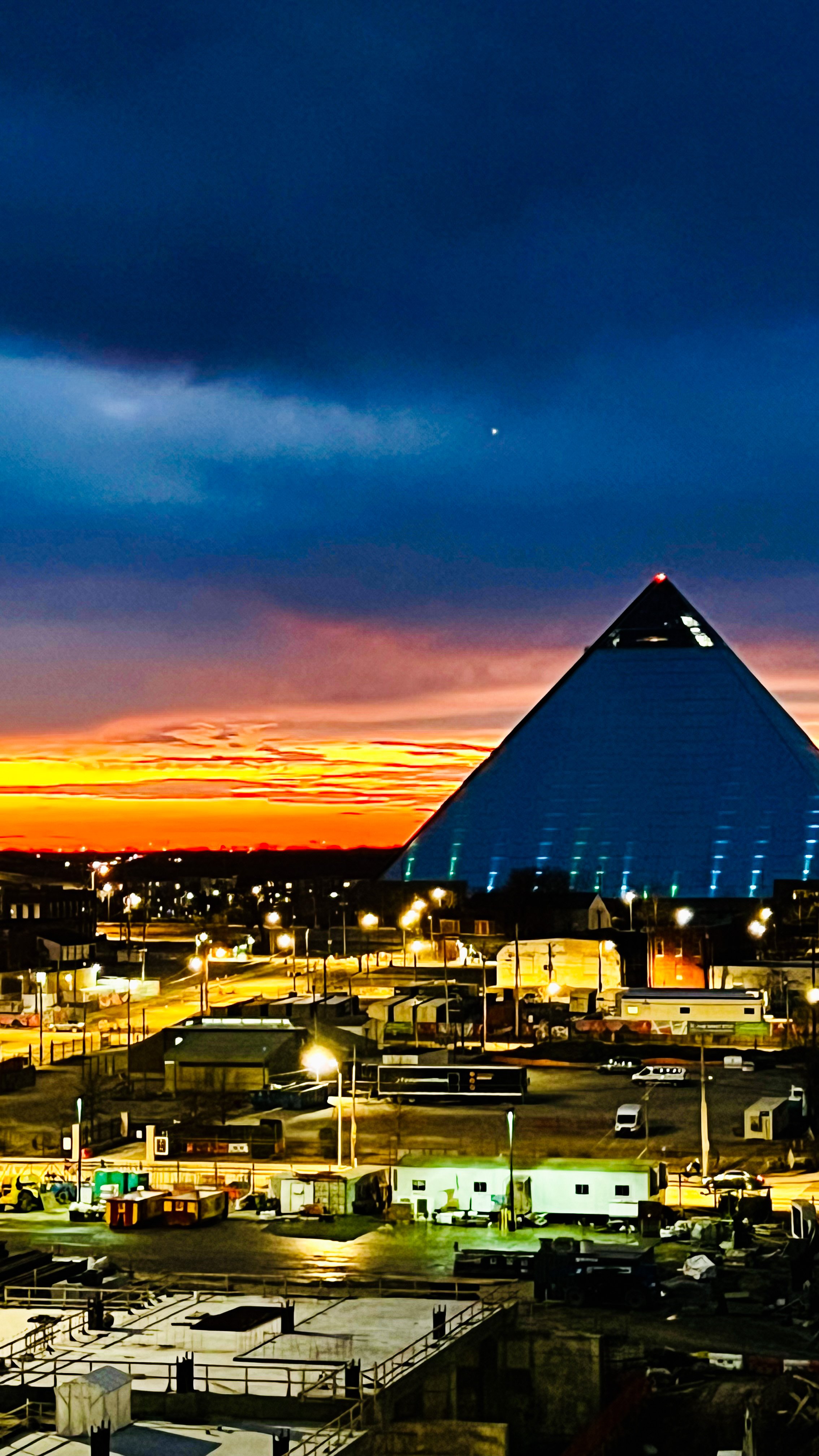 Nighttime cityscape with a large pyramid-shaped building illuminated against a colorful sunset sky.
