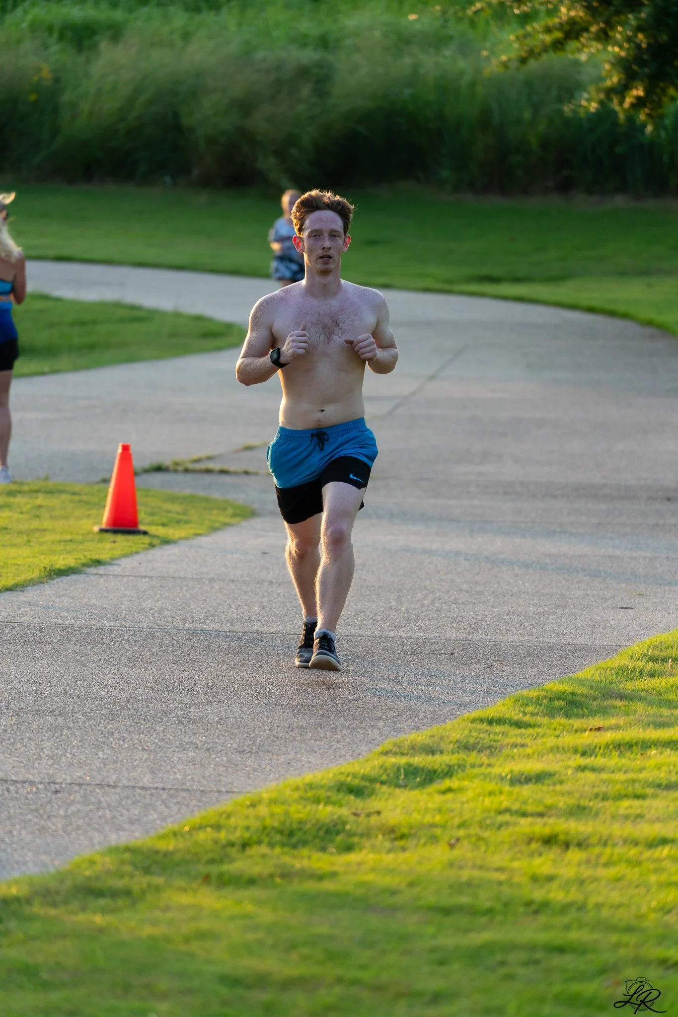 A shirtless man with red hair, running on a paved path in a park during sunset.