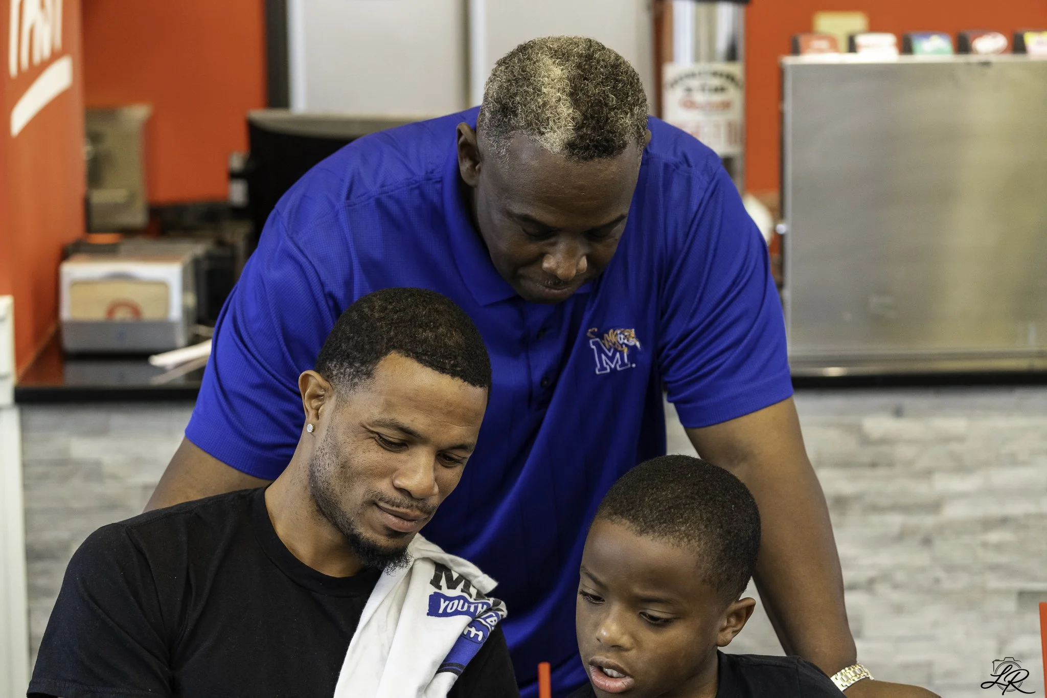Two young men sitting together and looking down, while a man in a blue shirt with a logo leans over them, in a casual setting with red accents and shelves in the background.