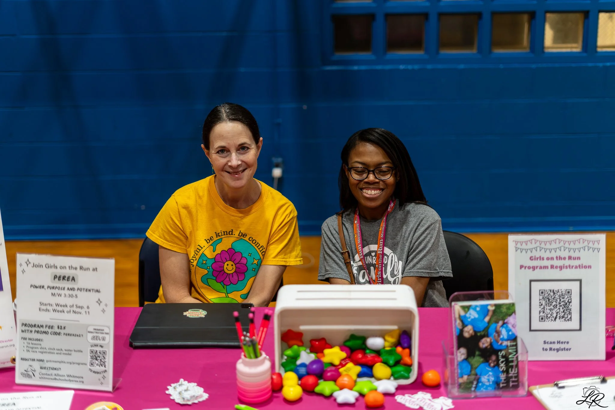 Two women are sitting behind a table at a registration booth, smiling. The table has colorful items and signs for the Girls on the Run program, with one sign indicating registration for girls and another with program details and a QR code.