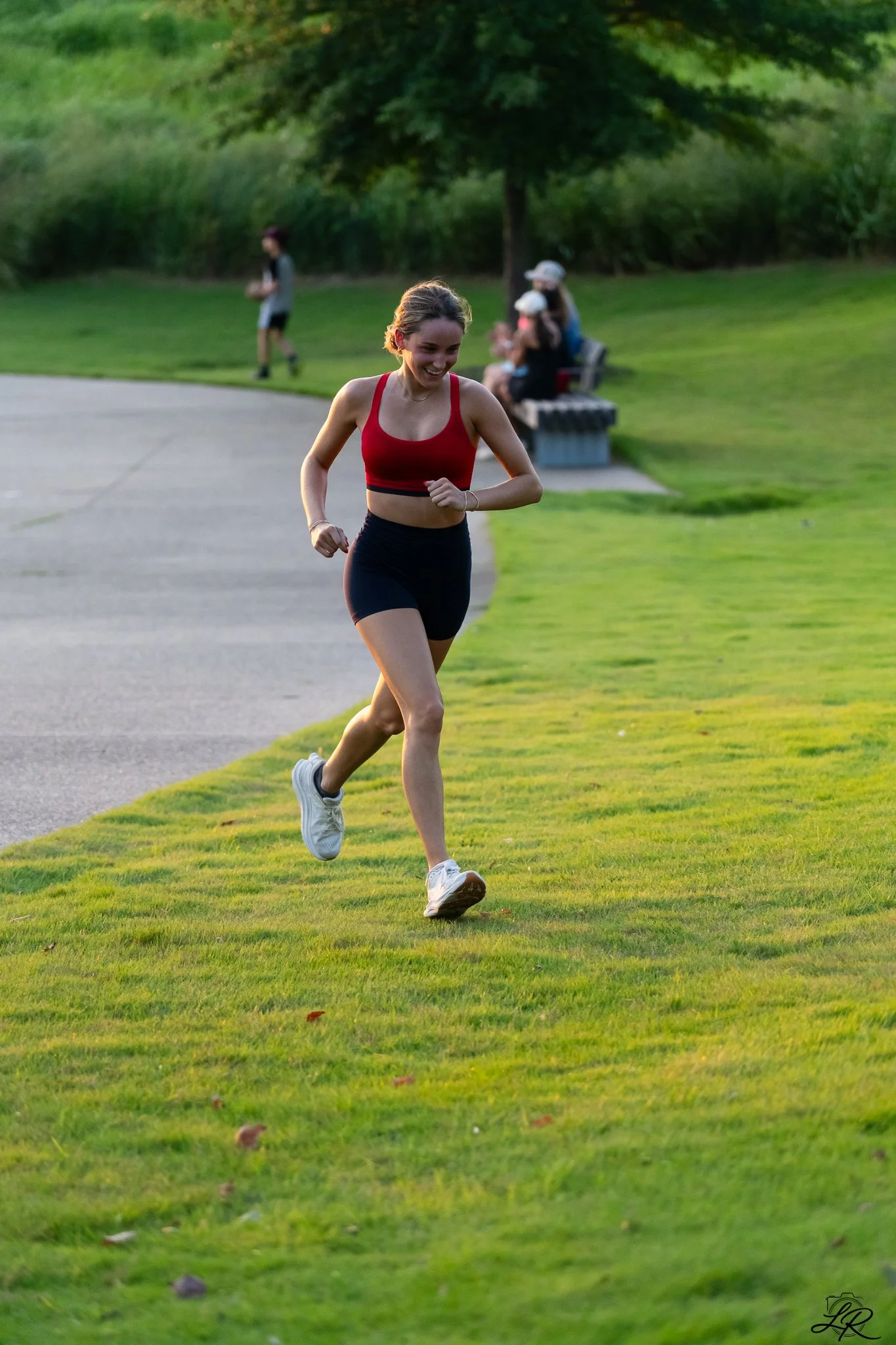 A young woman in athletic attire running on a grassy park path during late afternoon or early evening. She's smiling and appears to be enjoying her run, with trees and a park bench in the background.