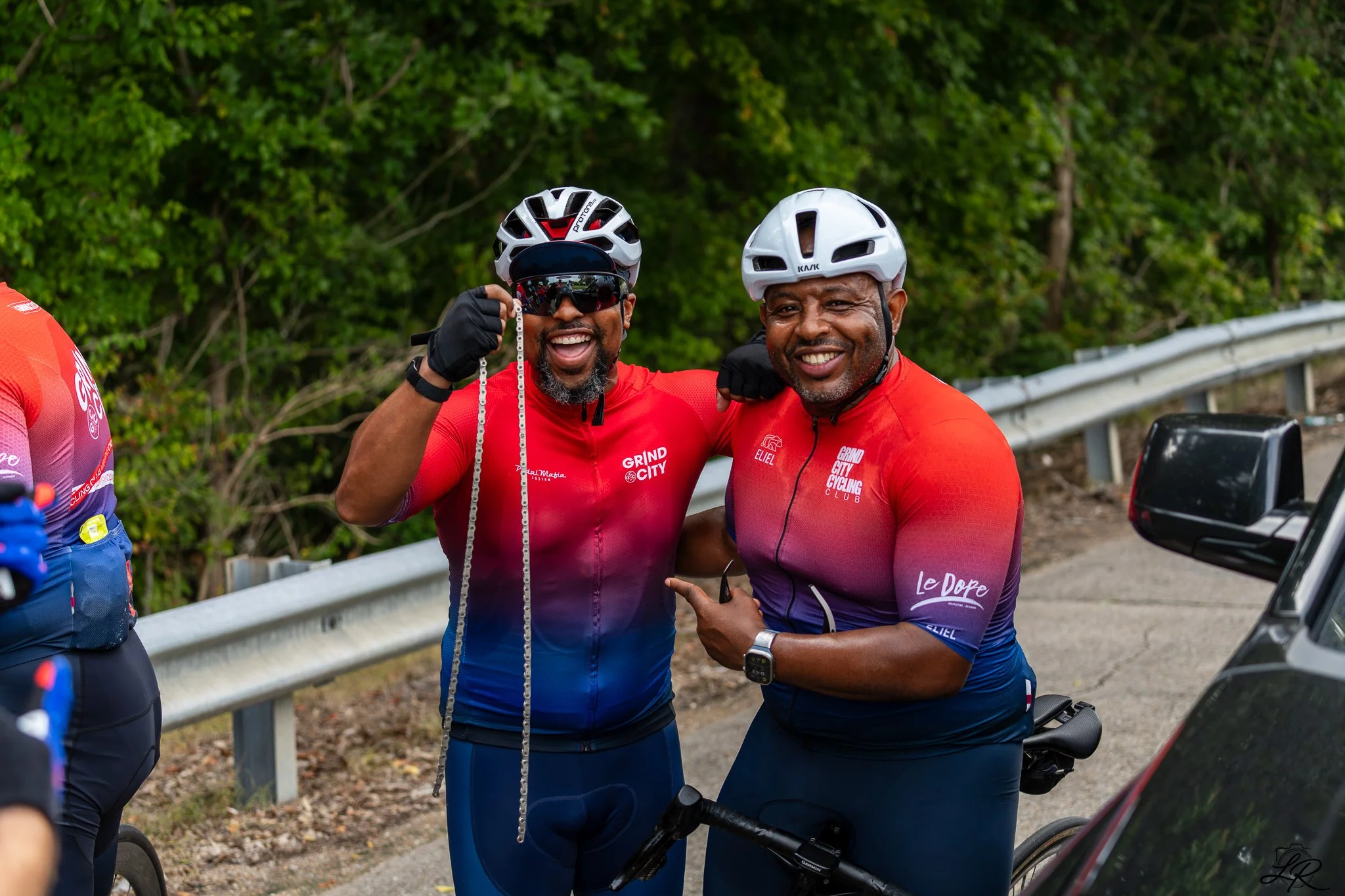 Two smiling cyclists wearing helmets and red cycling jerseys with logos, standing together outdoors near a metal guardrail and green trees, as one holds a necklace and the other points.