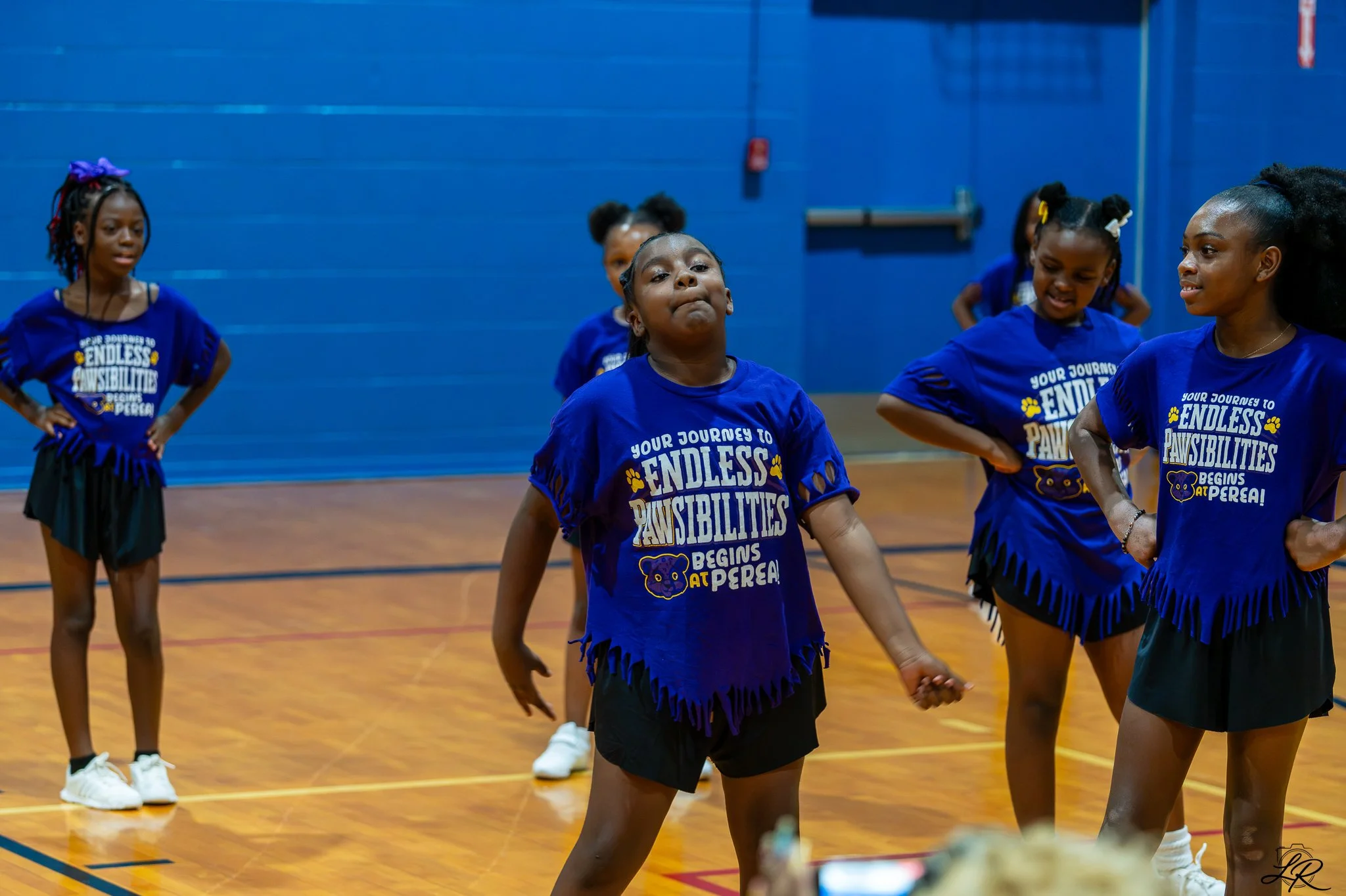 Group of young girls dancing in a gymnasium, wearing matching blue T-shirts with yellow text and black skirts, holding hands, with a blue wall and wooden floor in the background.