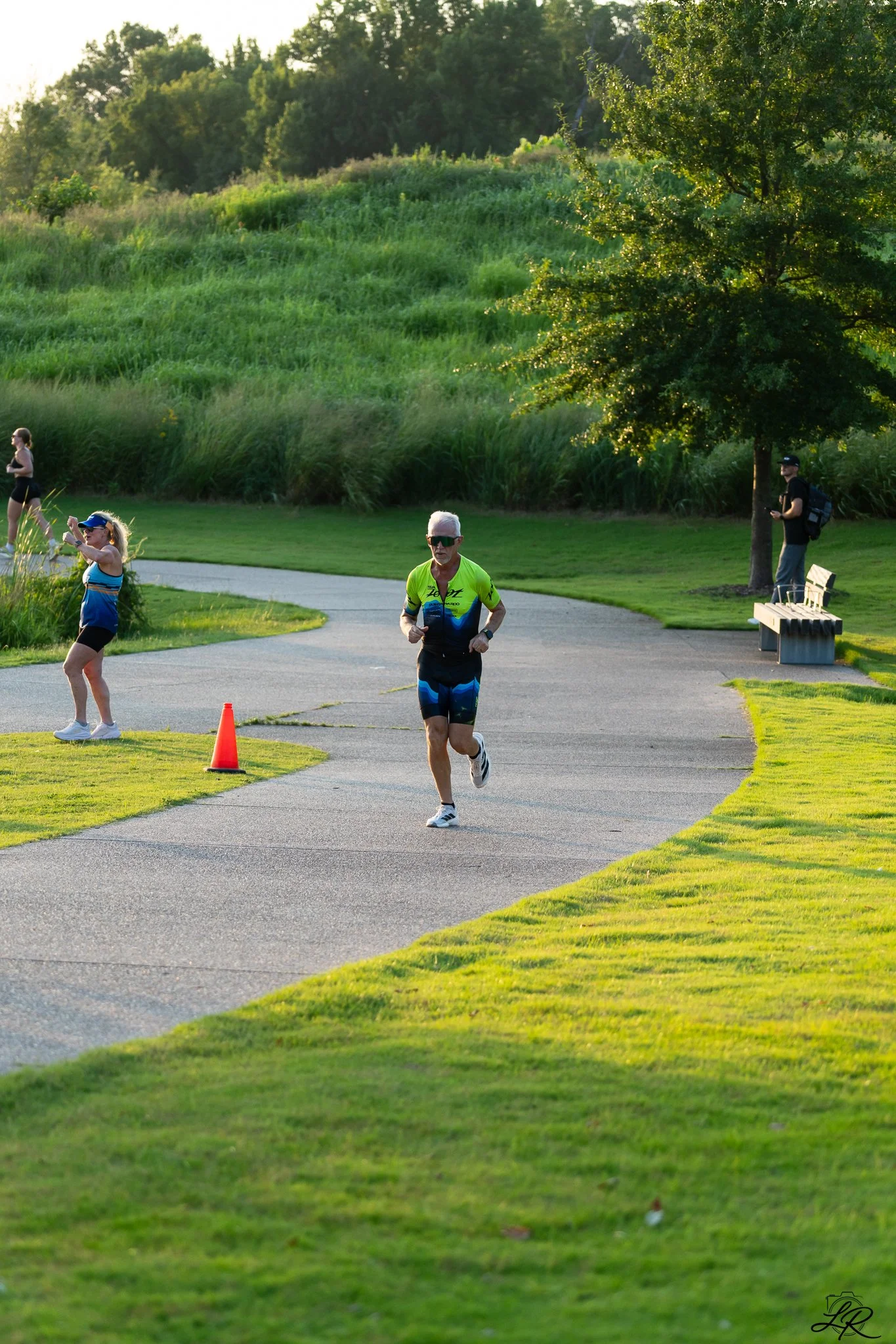 People running in a park or outdoor trail during the evening, with trees and green grass surrounding the paved path.