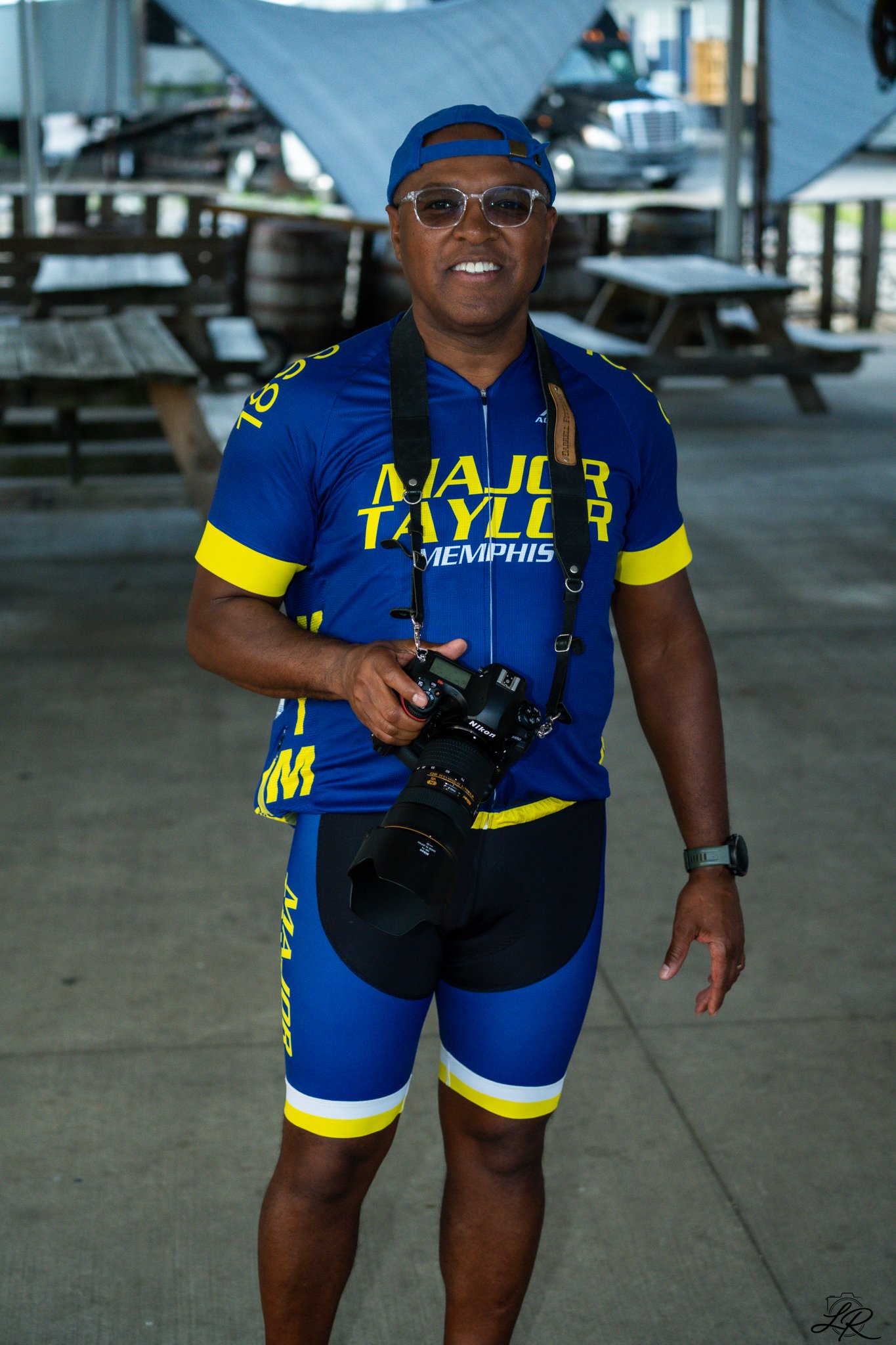 A man dressed in a blue and yellow cycling outfit, holding a camera, wearing glasses, a watch, and a blue cap, standing outdoors with benches, tables, and vehicles in the background.